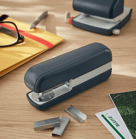 Kék tűzőgép fa asztalon, kapcsokkal, tollal és papírokkal körülvéve.