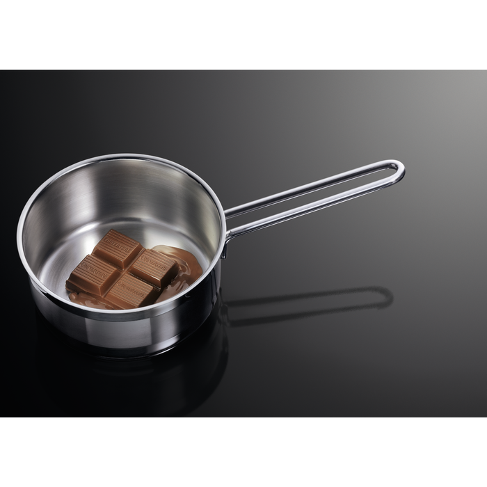 Cacerola de acero inoxidable con cuadrados de chocolate derritiéndose dentro. Está sobre una superficie negra, con un reflejo oscuro.