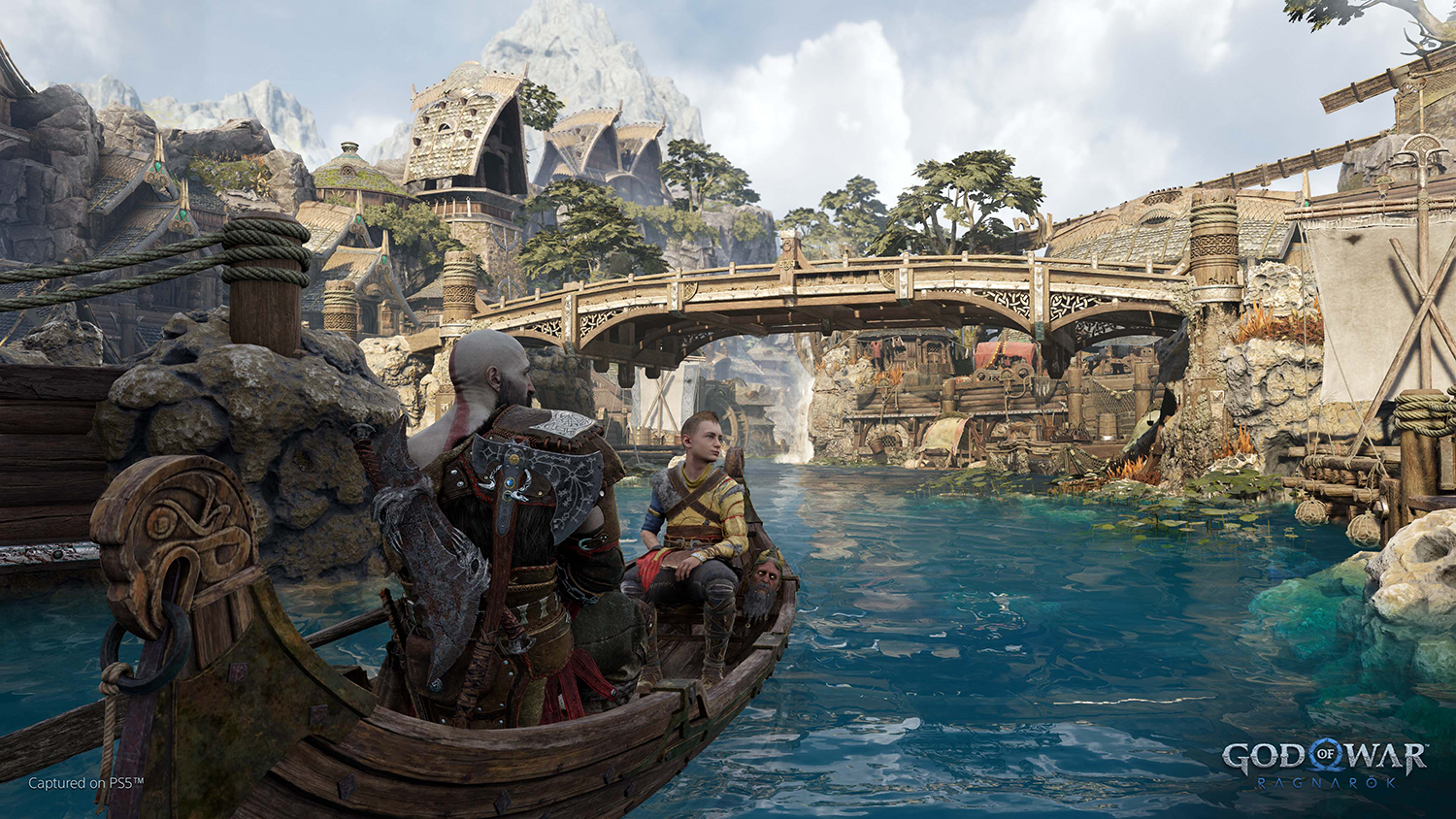 Two people in a boat, sailing on a lake. Mountains and a bridge in the background.