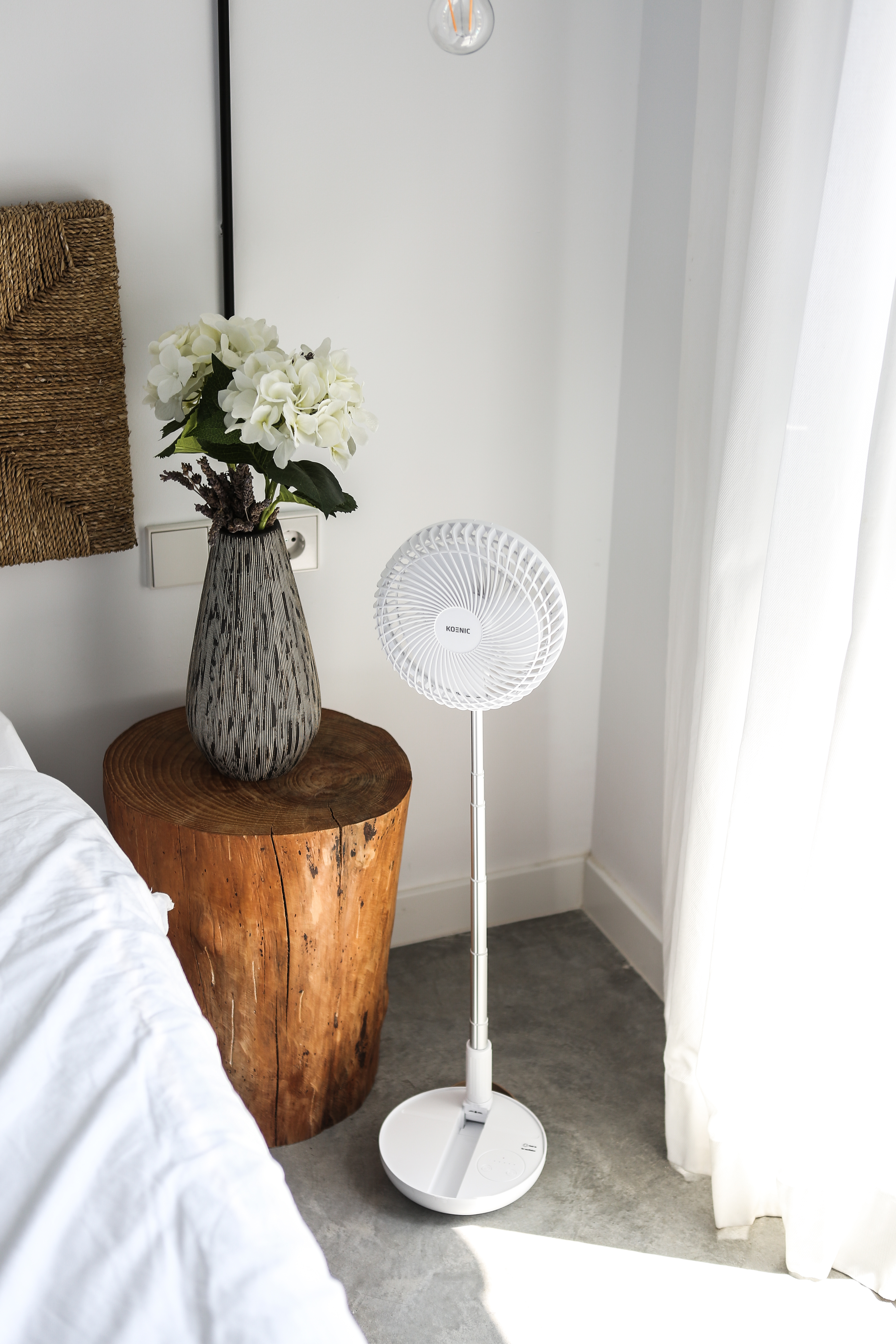 Fehér ventilátor fehér állványon, világos padlón. Egy vázában virágok egy fa kisasztalon.
