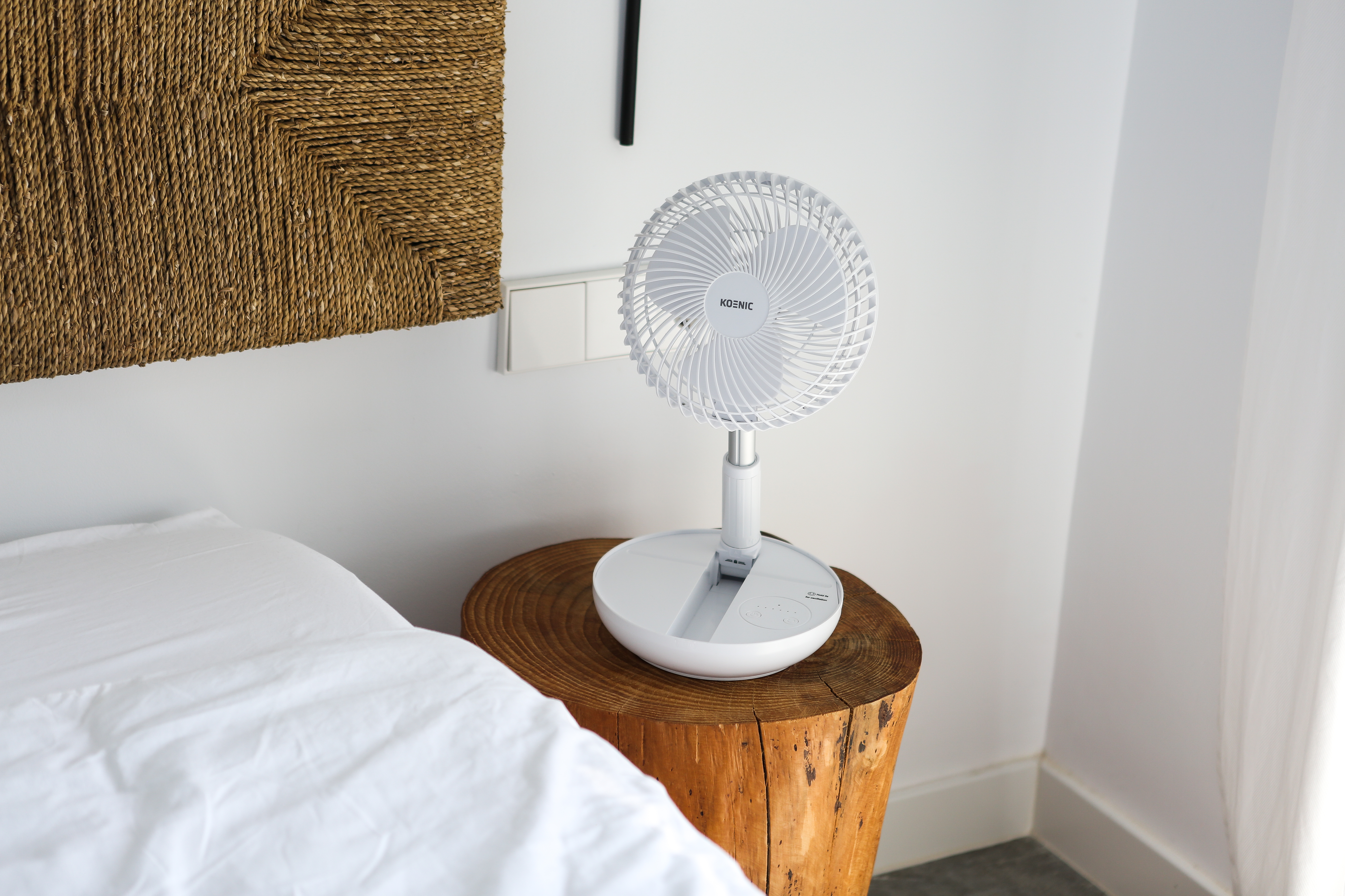 Egy fehér asztali ventilátor egy fa csonkon, egy fehér lepedős ágy mellett.