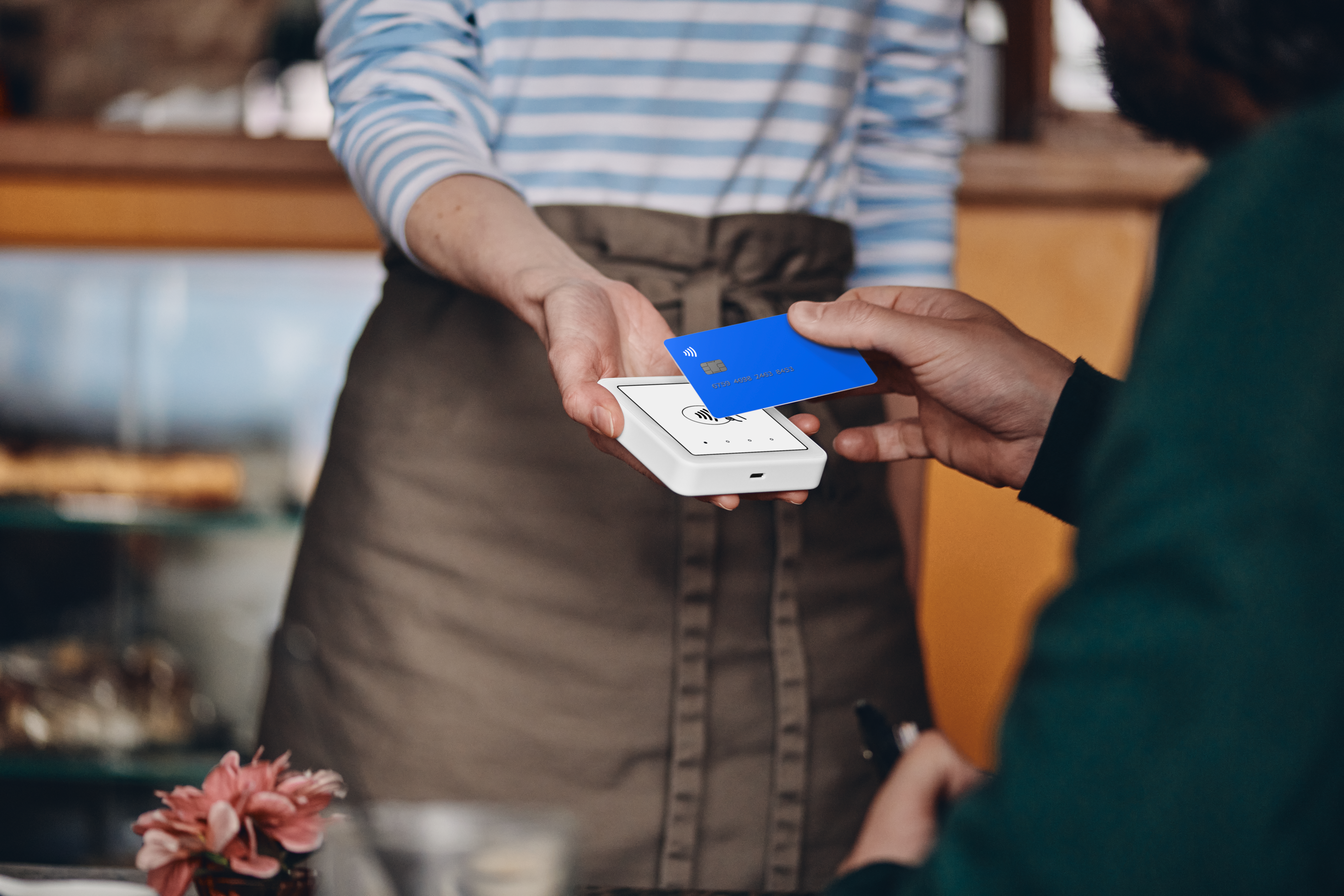 Eine Person gibt eine blaue Kreditkarte an eine andere Person über ein Zahlungsterminal in einem Café.