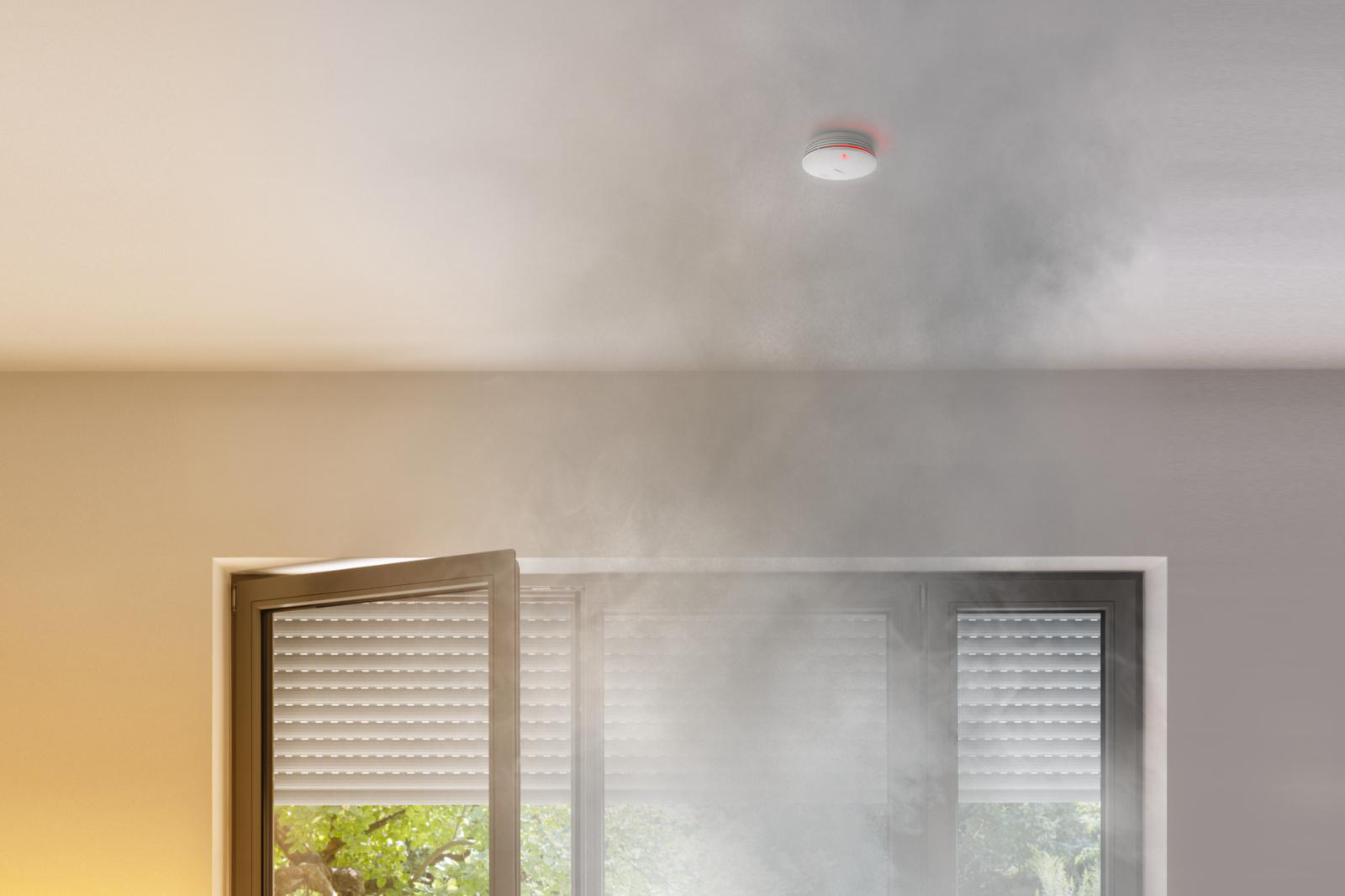 Rauch füllt einen Raum mit einem Rauchmelder an der Decke. Ein offenes Fenster befindet sich im Vordergrund.