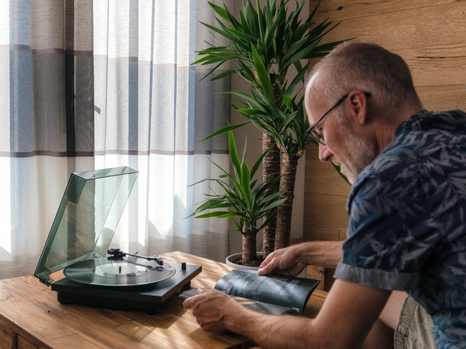 Un homme avec des lunettes est assis, regardant la pochette d'un album, à côté d'une platine et d'une plante.