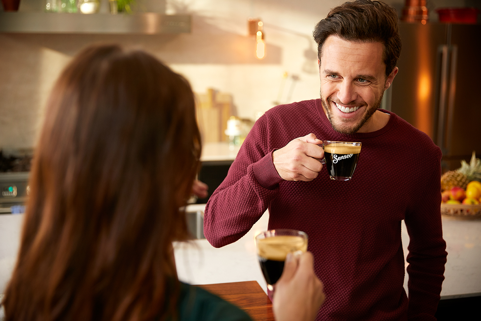 A man and woman in a kitchen, drinking coffee. Smiling man, coffee cups visible.