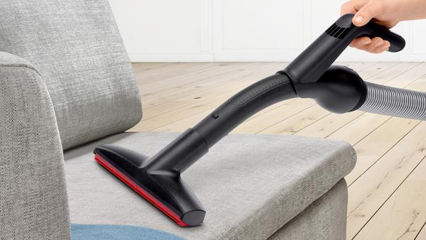 Eine Hand saugt einen grauen Sofa ab. Der Staubsaugerkopf ist schwarz mit rotem Rand.