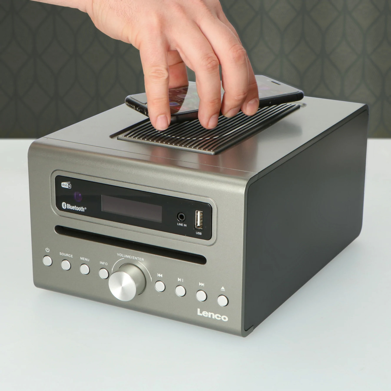 Une main posant un téléphone sur une radio Lenco grise. La radio a des boutons, un écran et une fente CD.