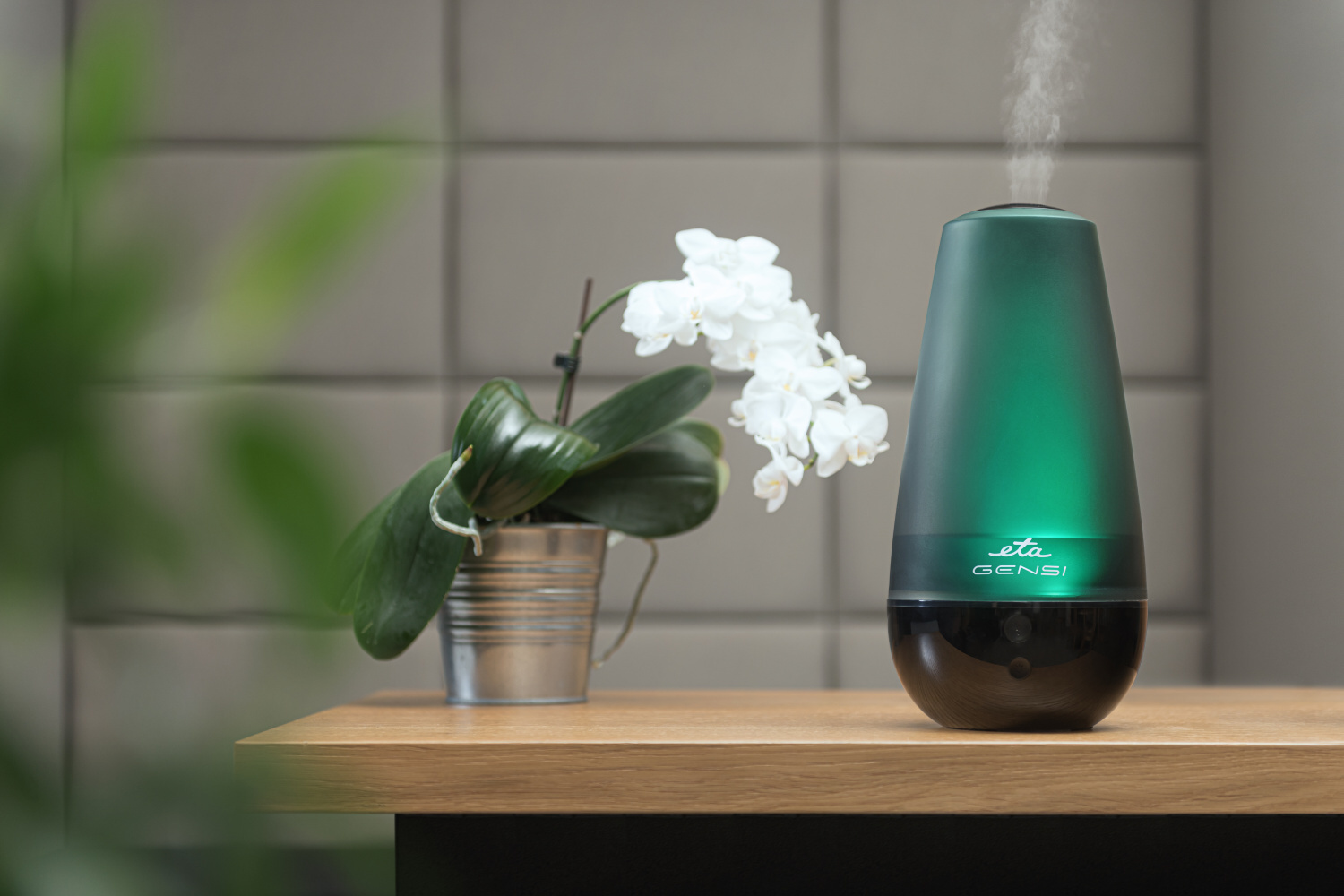 Aromadiffúzor zöld ragyogással, fehér virágokkal és egy ezüst cserépben lévő növénnyel a faasztalon.
