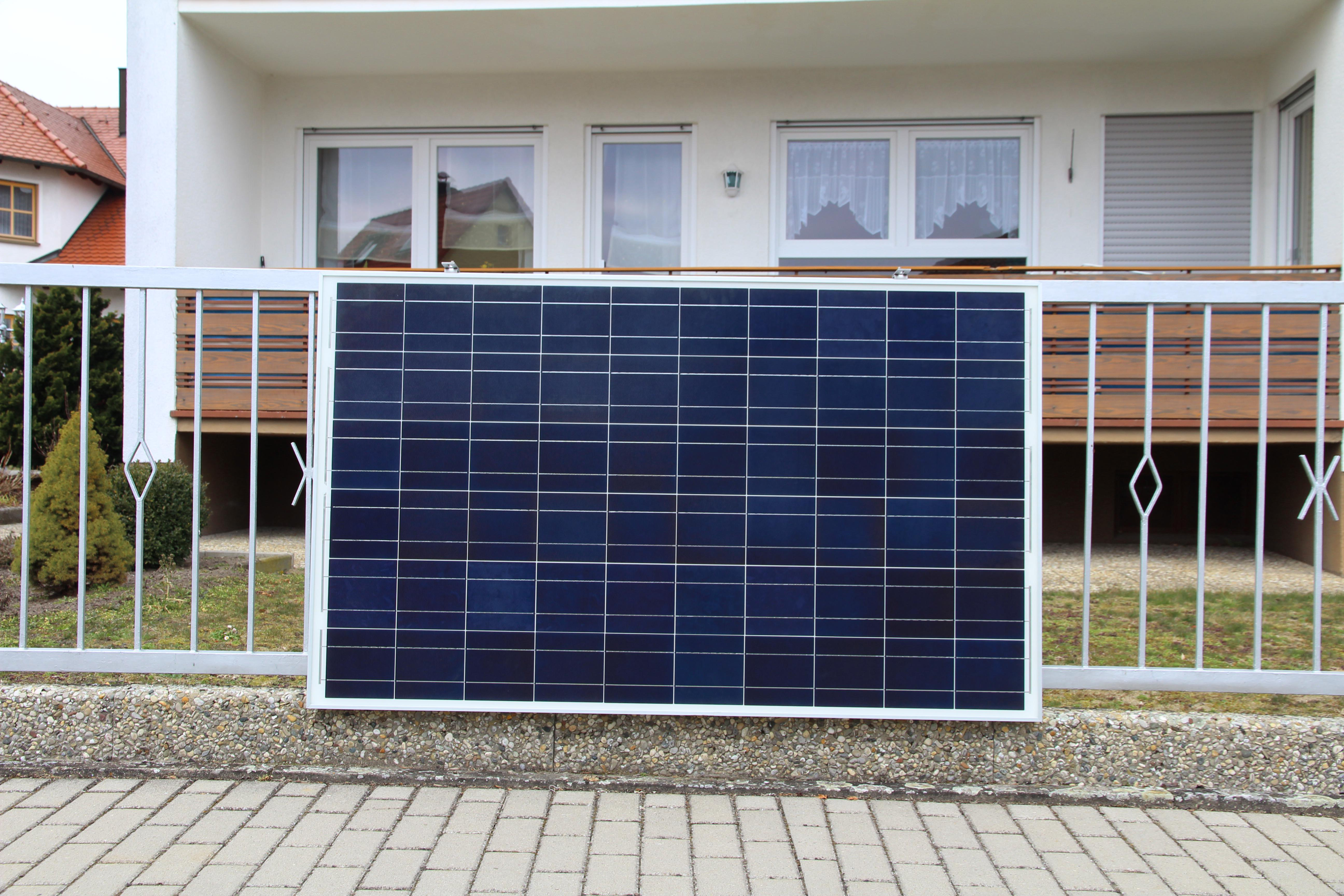 Ein Solarpanel, das an einem Metallzaun befestigt ist. Ein Haus ist hinter dem Zaun sichtbar.