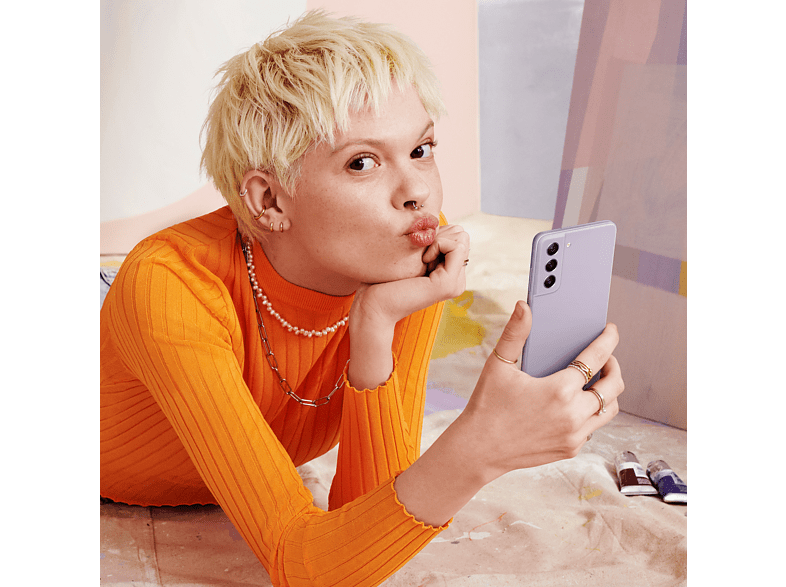 Mujer con pelo corto rubio, top naranja, sosteniendo teléfono lila, haciendo pucheros. Pinturas al frente.
