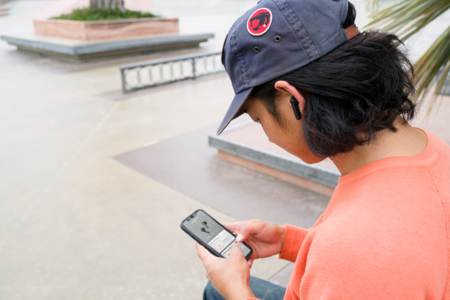 Man met een pet en oortelefoons, kijkt naar een telefoon. Vage achtergrond van een skatepark.