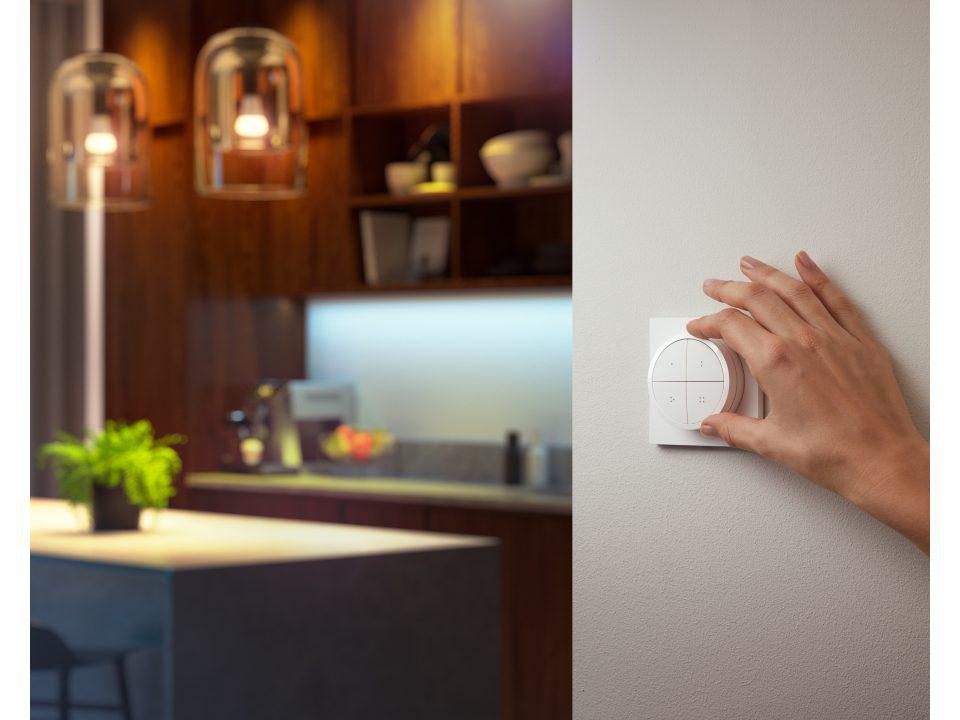 Hand dreht einen weißen Smart-Lichtschalter an einer Wand, im Hintergrund eine Küche.