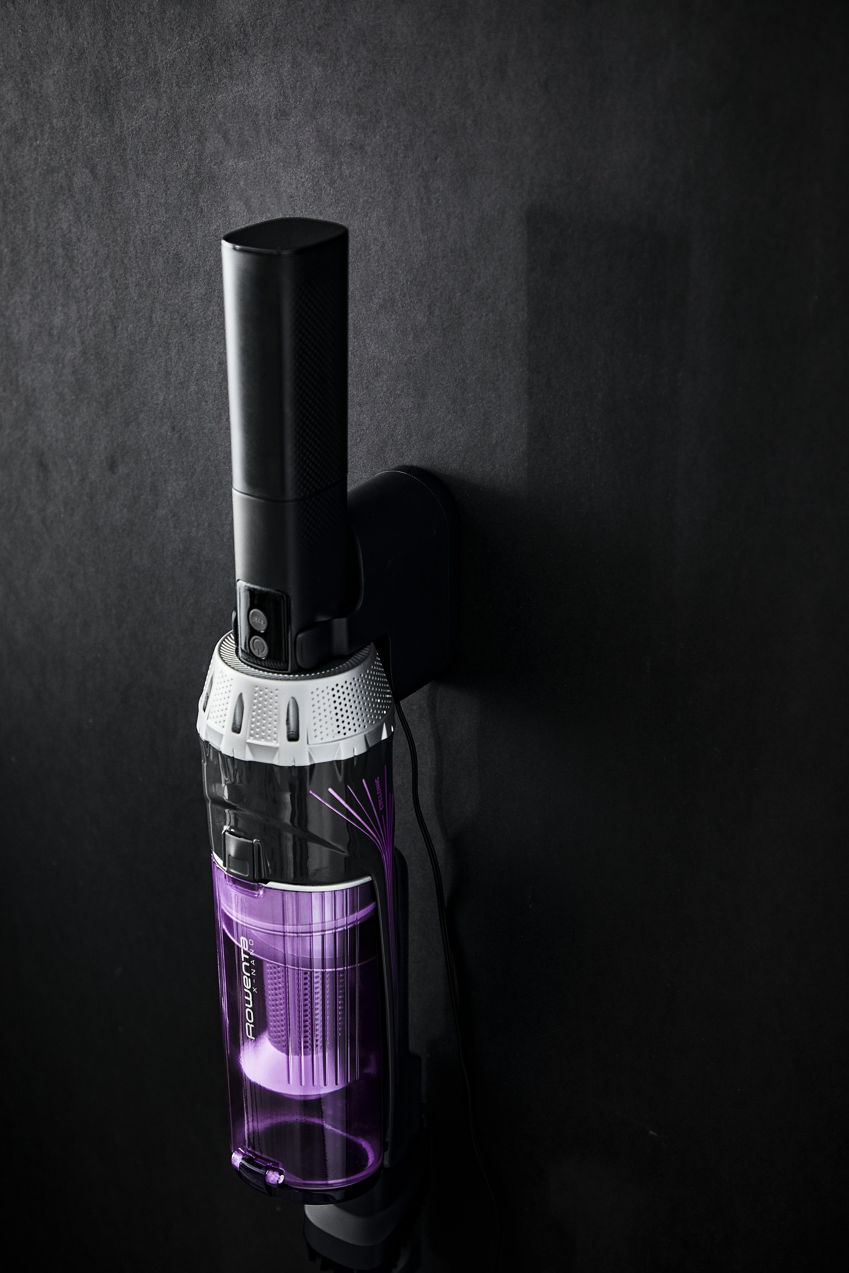 Ein Staubsauger ist an einer dunklen Wand montiert. Der transparente Staubbehälter zeigt einen violetten Innenraum.