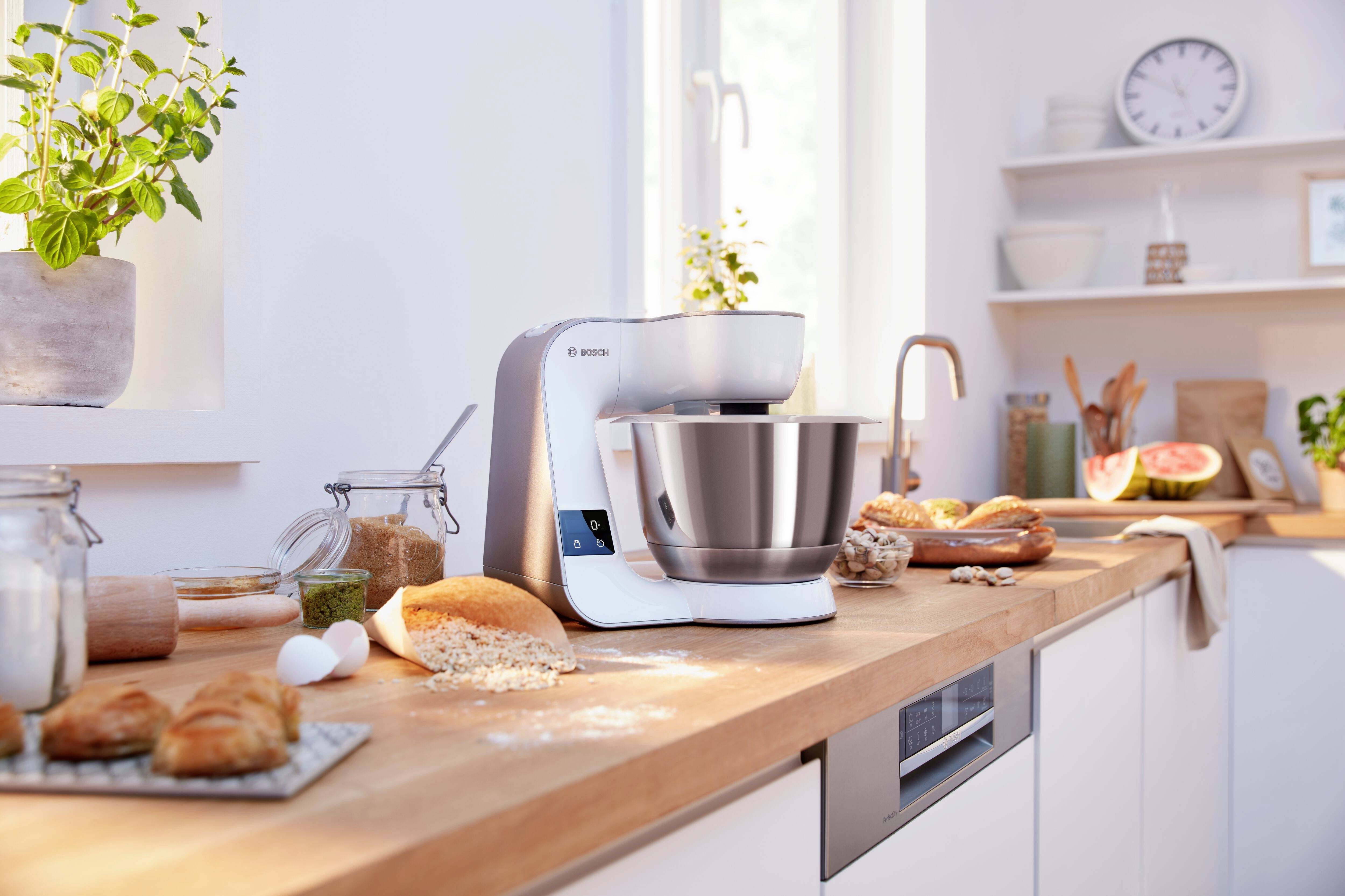 Una cocina con una batidora blanca sobre una encimera de madera. Hay pan, huevos y una planta cerca. Hay una ventana detrás.