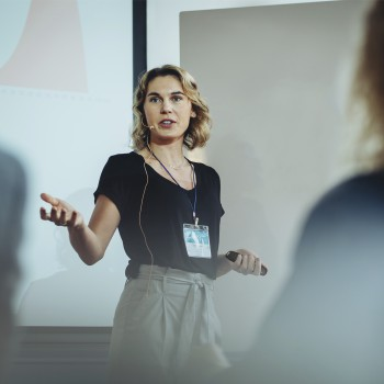 Una mujer está dando una presentación en un escenario. Gesticula con ambas manos. Una pantalla está detrás de ella.