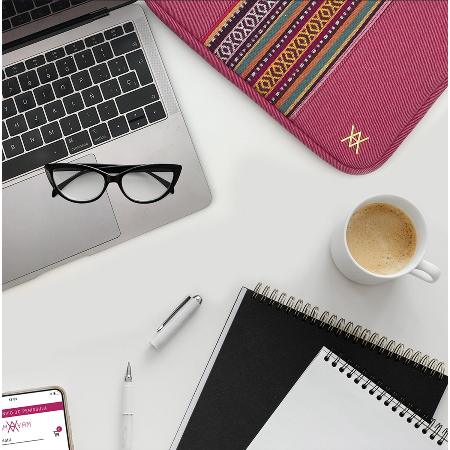 Espacio de trabajo con portátil, gafas, cuaderno, bolígrafo y taza de café sobre una superficie blanca.