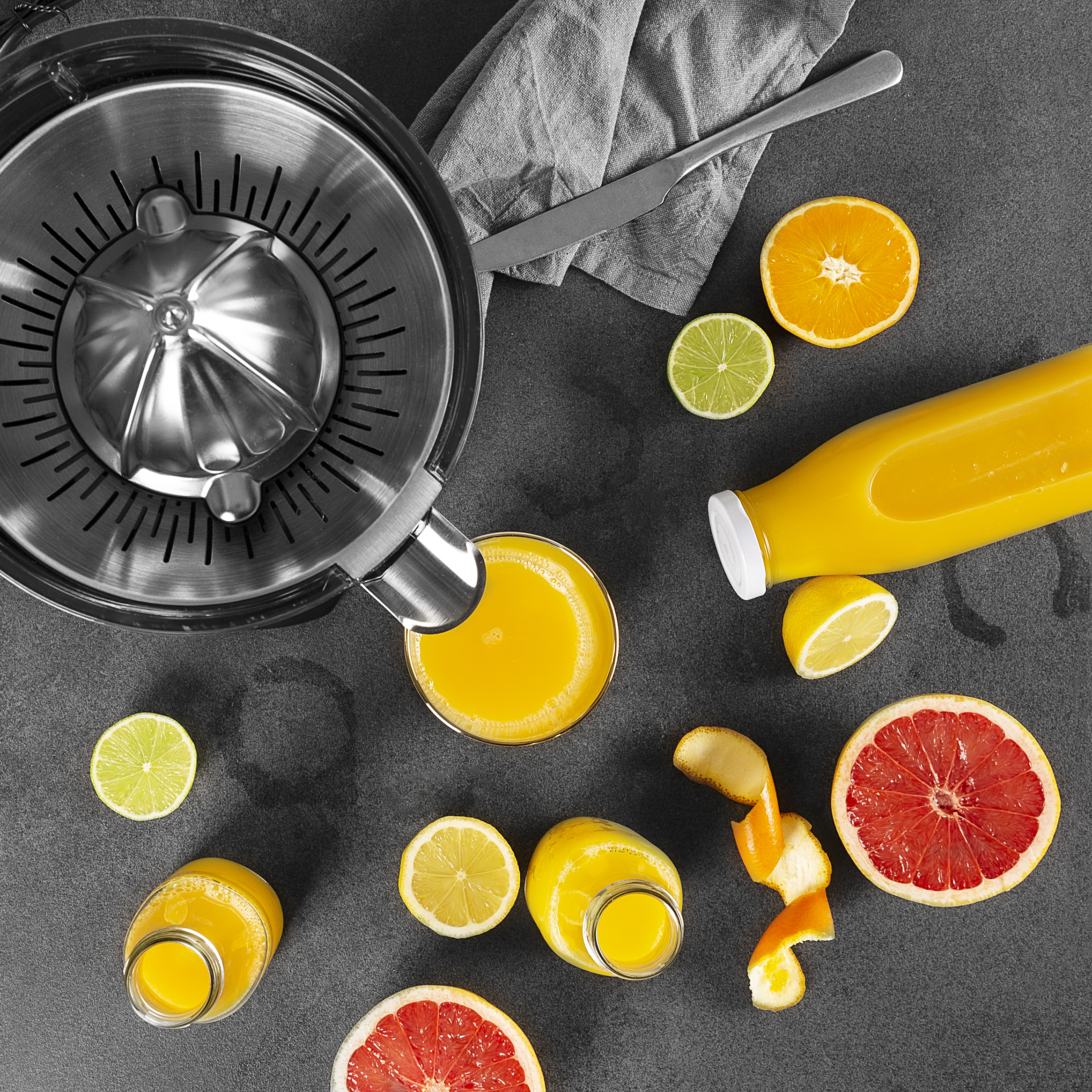 Ophangfoto van een citruspers, sapflessen, citrusvruchten en een mes op een donkergrijze ondergrond.