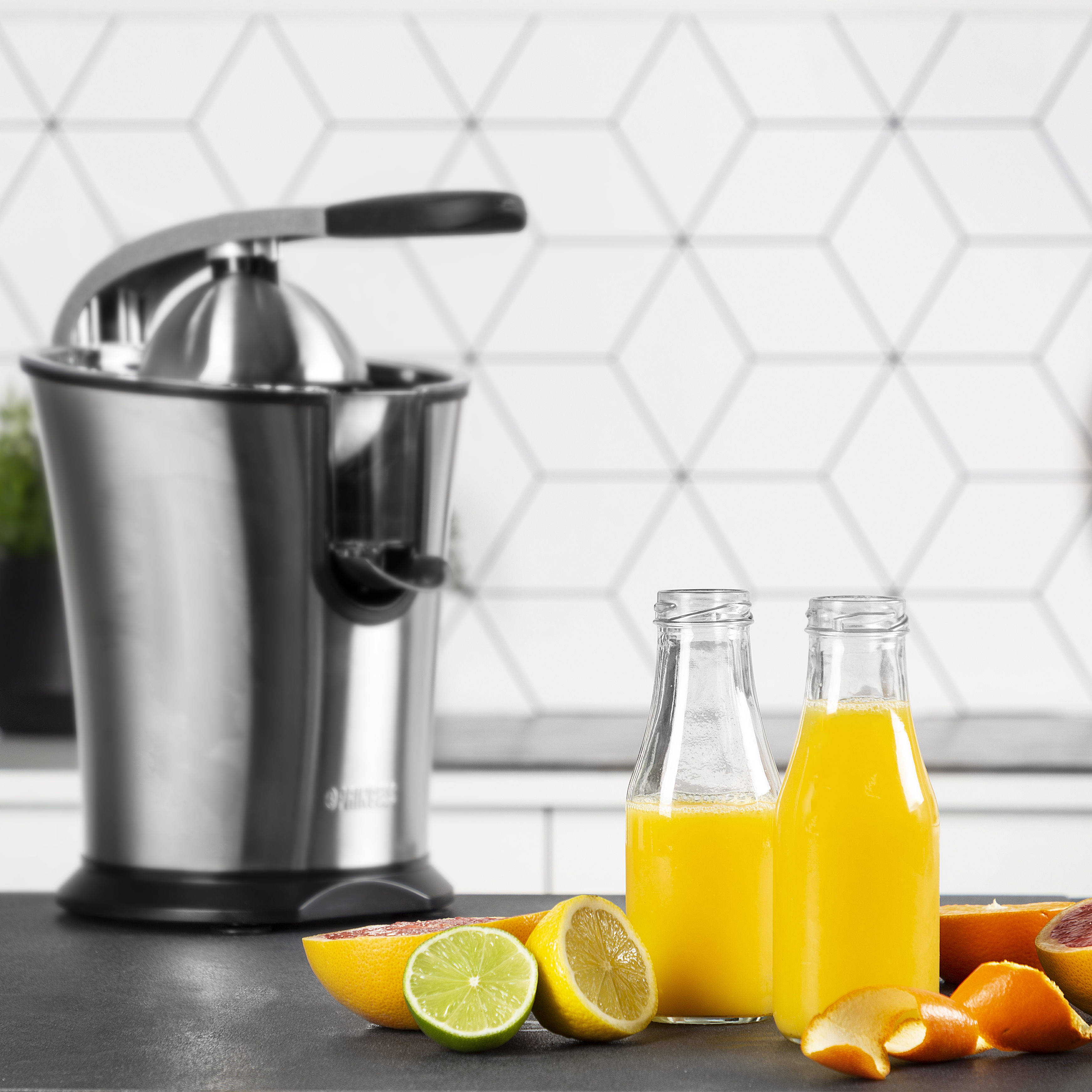 Een roestvrijstalen juicer met twee sapflessen, citroenen en sinaasappels op een donker oppervlak.