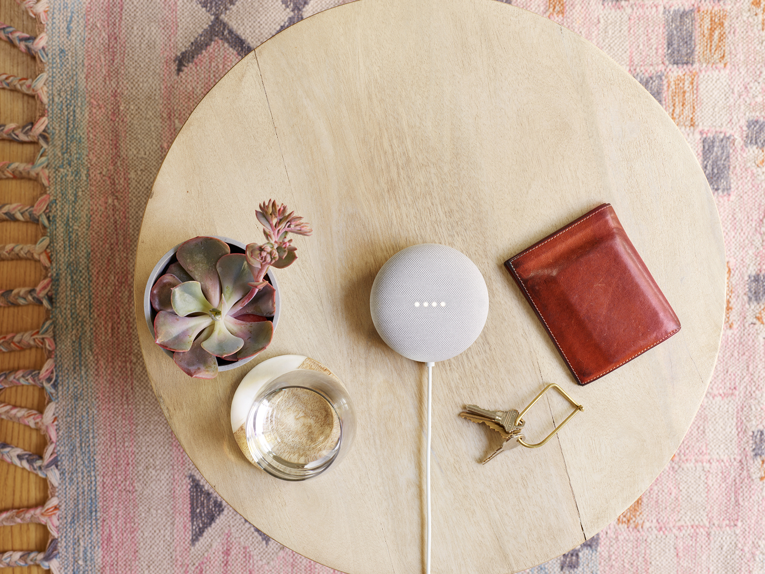 Houten tafel met Google Nest Mini, vetplant, portemonnee, sleutels en glas. Vloerkleed zichtbaar.