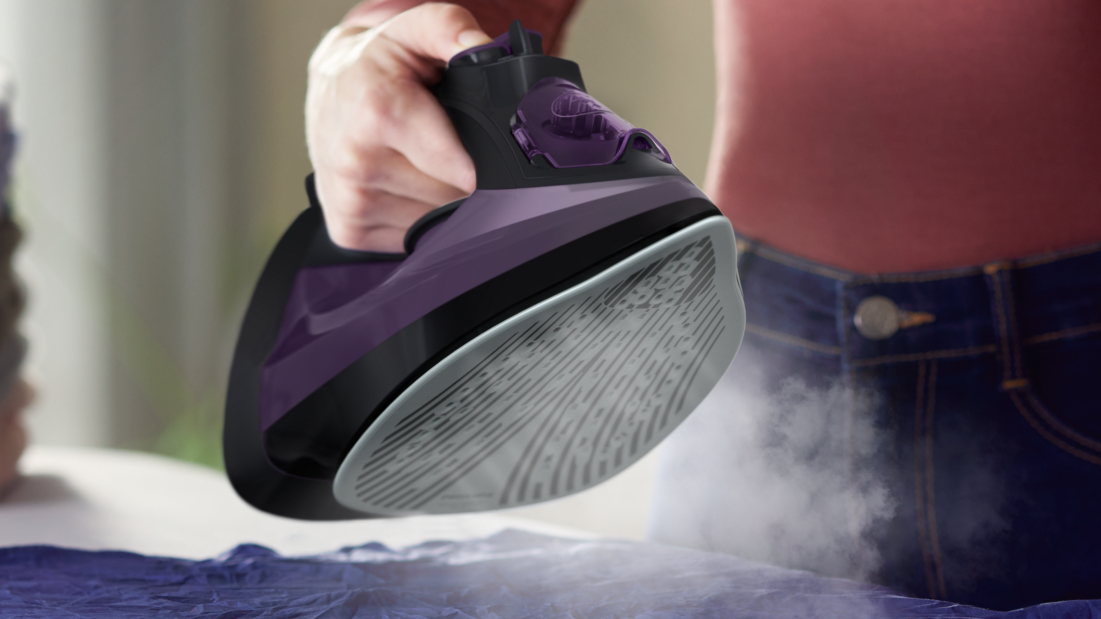 Una plancha morada y negra vaporizando tela. La mano de una persona sostiene la plancha.