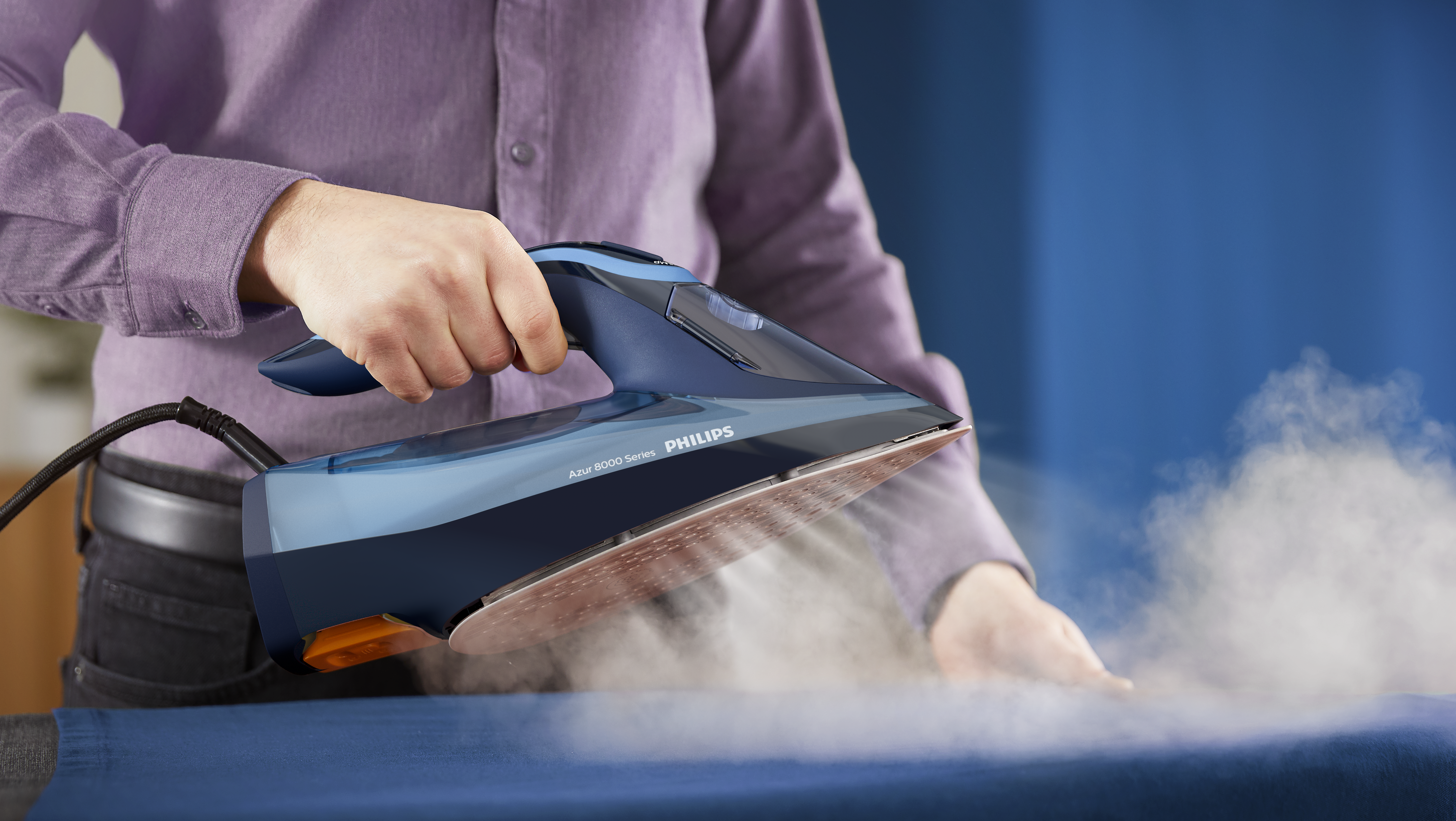Persona planchando tela azul con plancha Philips, vapor subiendo. Camisa morada al fondo.