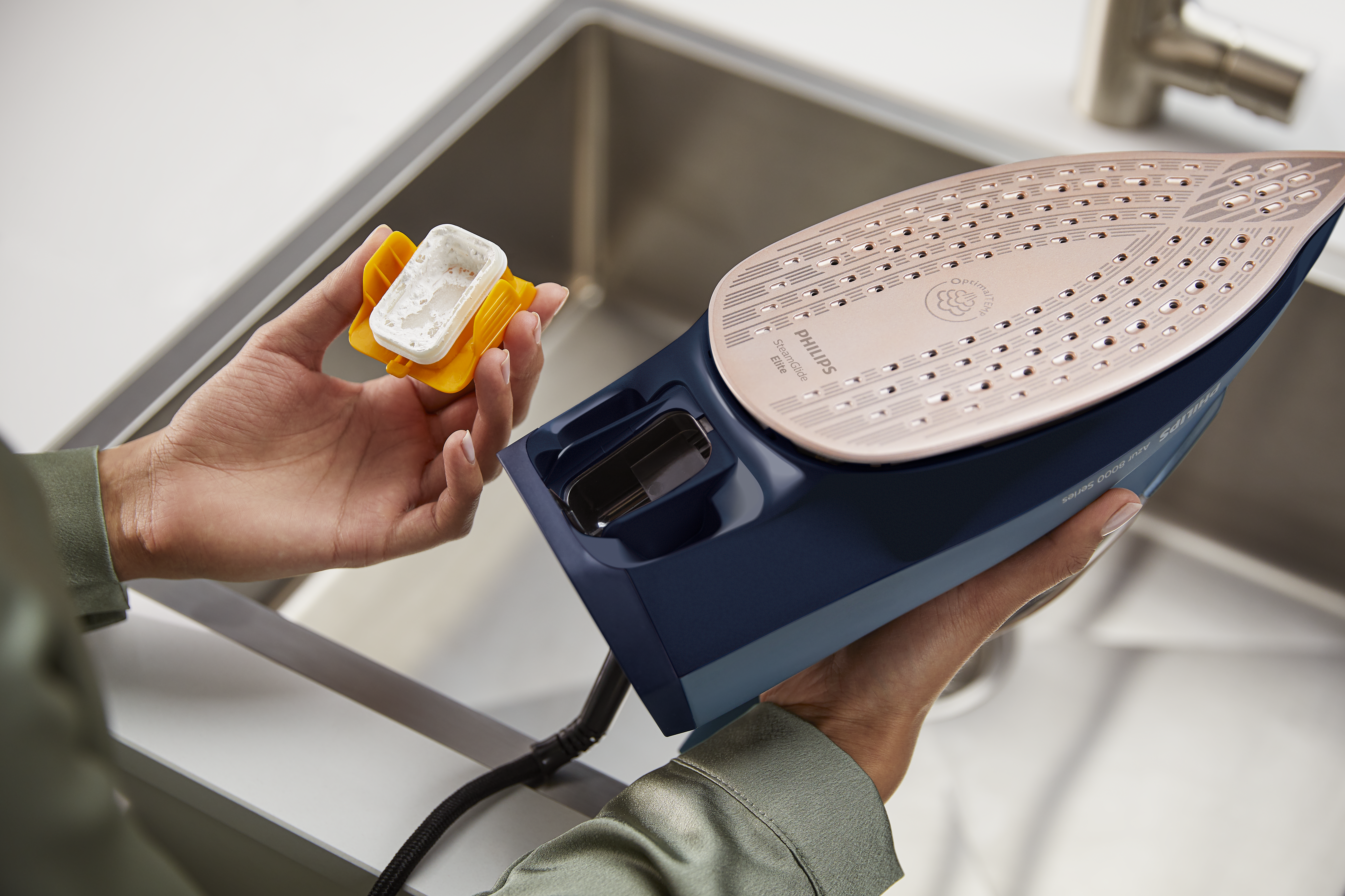 Una persona sostiene una plancha Philips y un cartucho descalcificador. La plancha es azul y dorada, cerca de un fregadero.