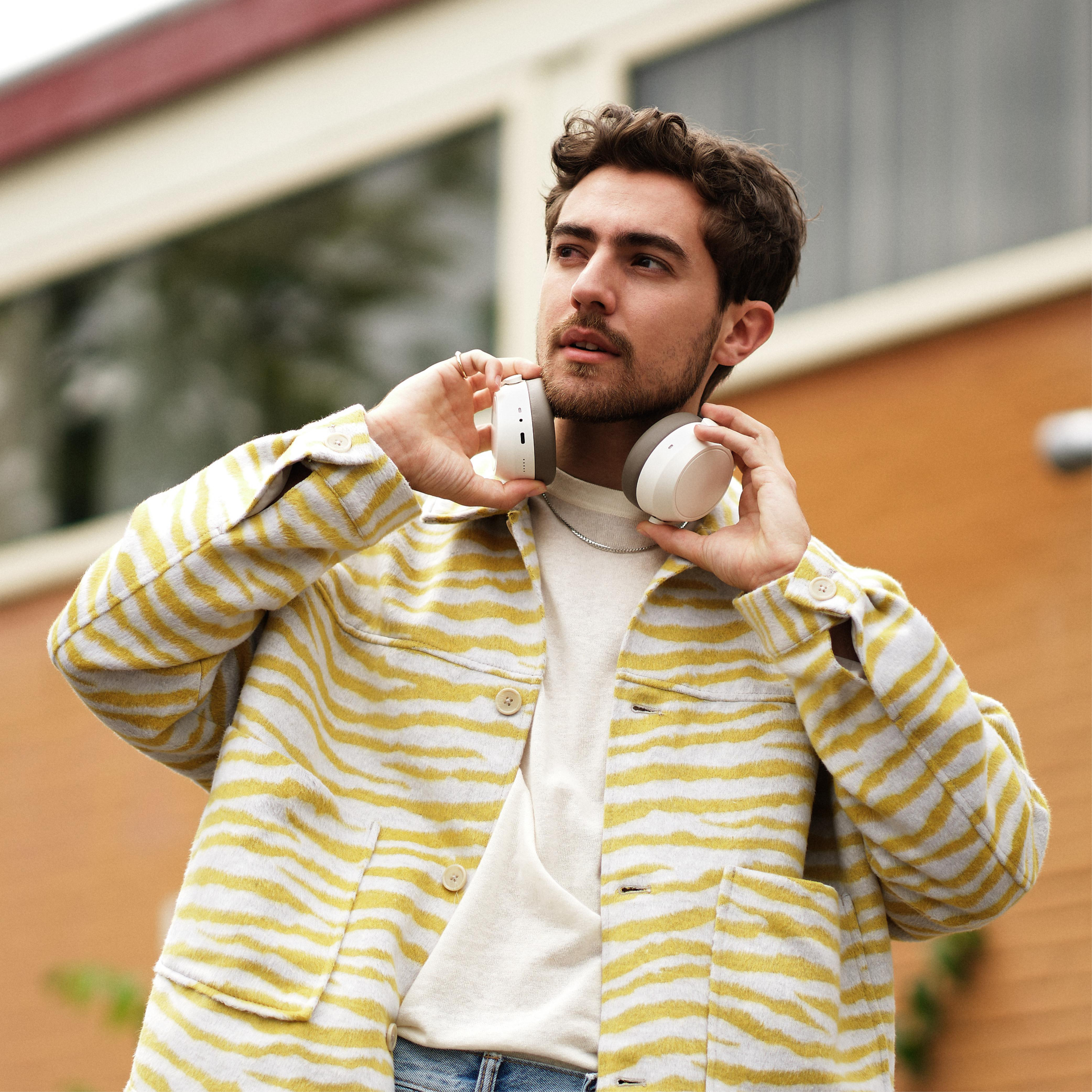 Mann hält weiße Kopfhörer, trägt eine gelb-weiß gemusterte Jacke.