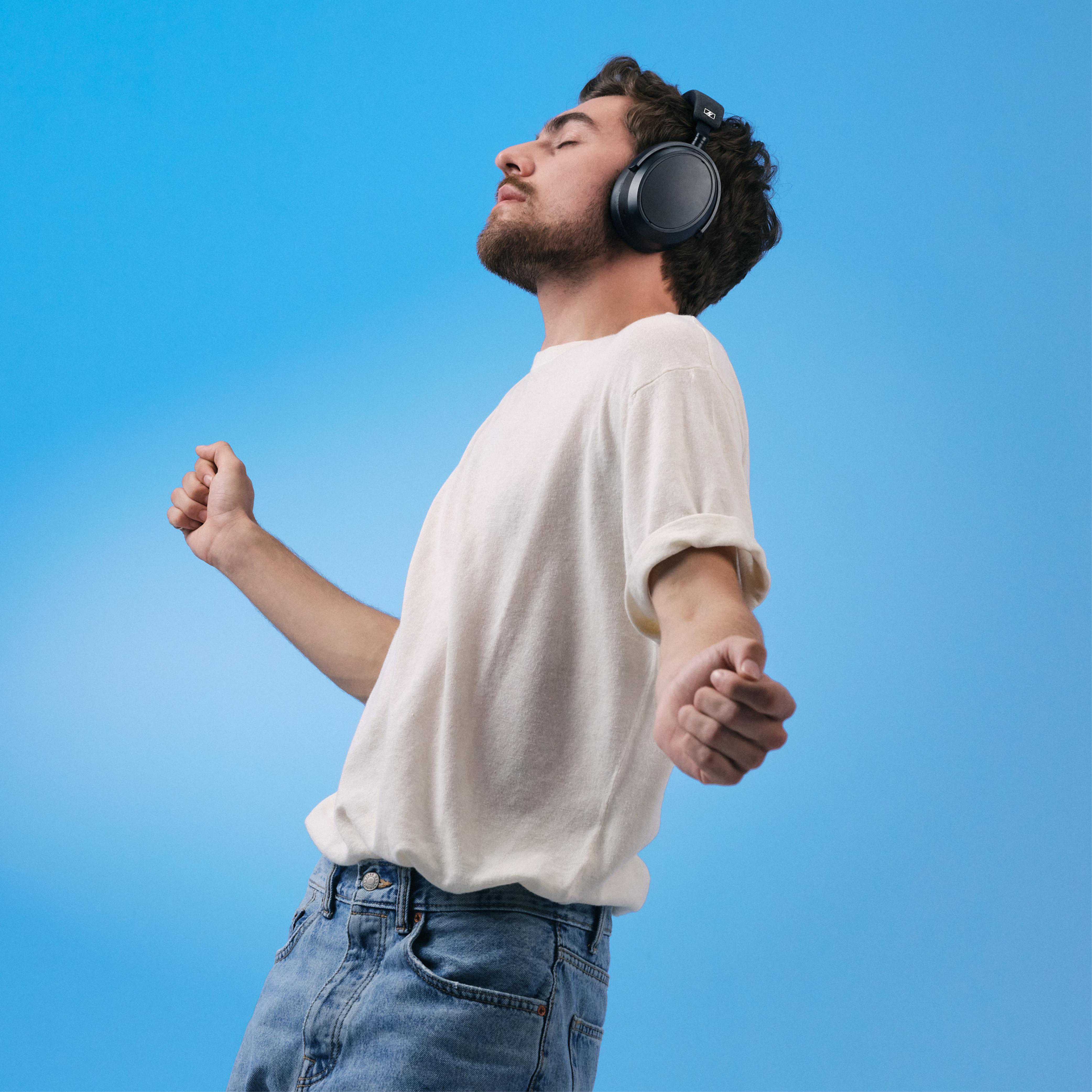 Mann mit Kopfhörern, Arme ausgestreckt, Augen geschlossen, vor blauem Hintergrund.