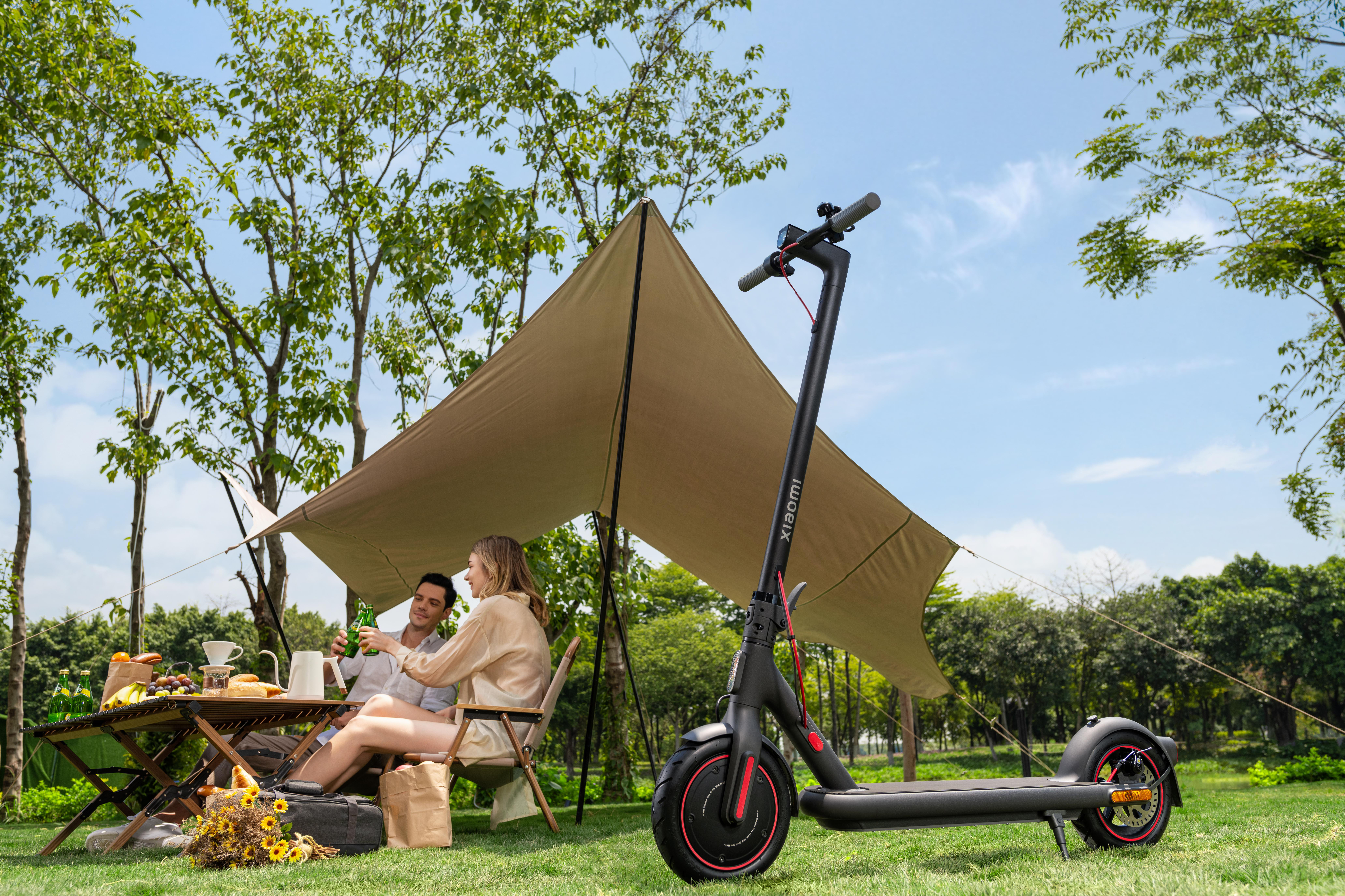 Eine Frau und ein Mann sitzen an einem Tisch neben einem Roller in einem Park mit Bäumen und einem Zelt.