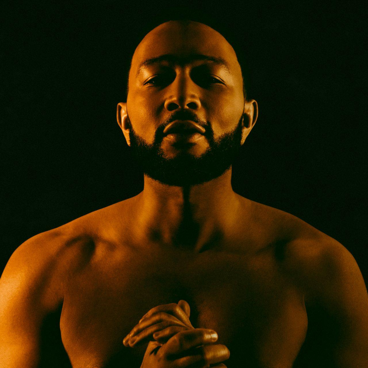 John Legend torse nu, mains jointes, face au spectateur. Fond sombre.