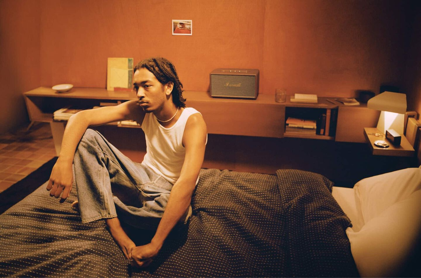 Ein Mann in einem weißen Tanktop sitzt auf einem Bett in einem Raum mit einem Marshall-Lautsprecher.
