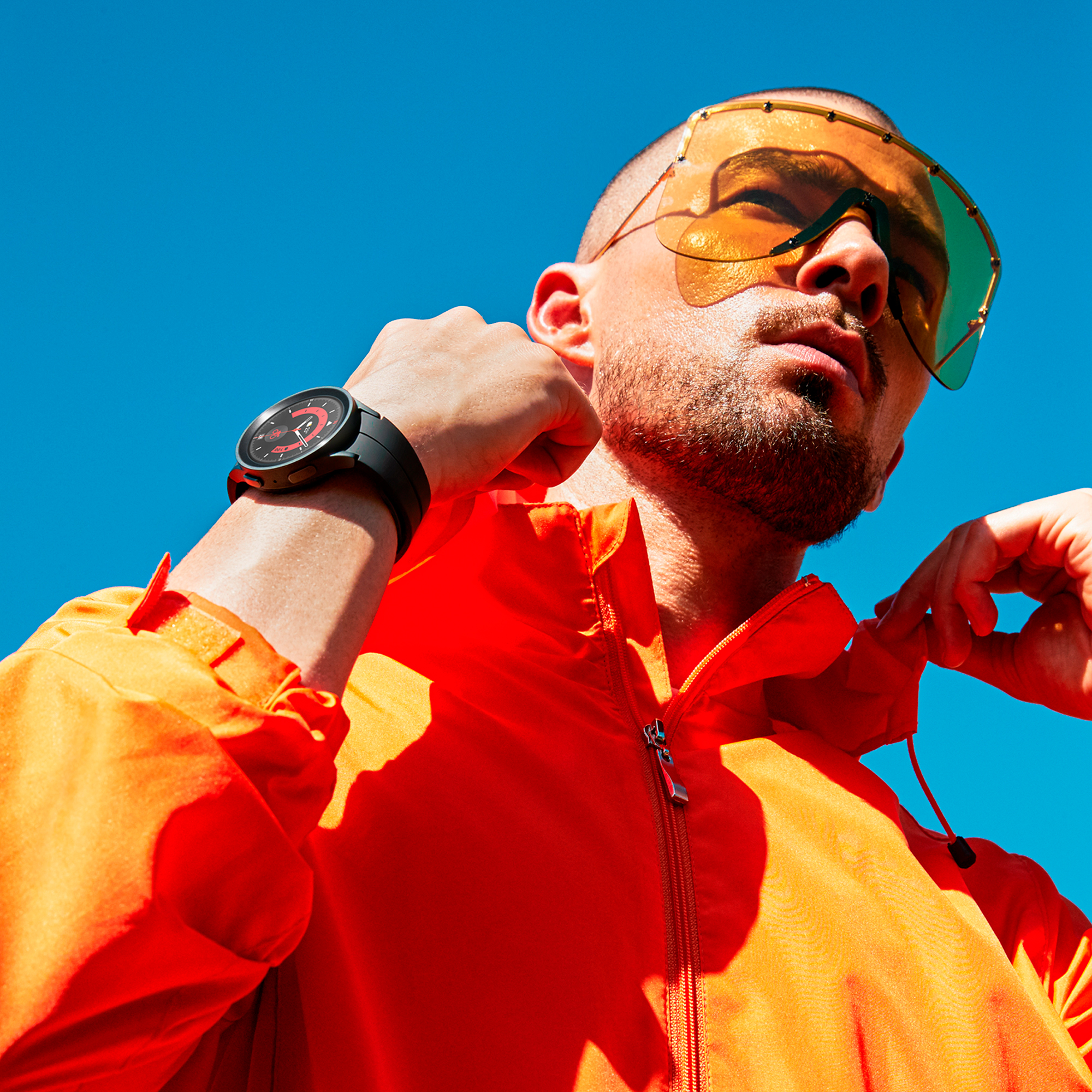 Hombre con chaqueta naranja y gafas de sol amarillas usando reloj negro, fondo de cielo azul.