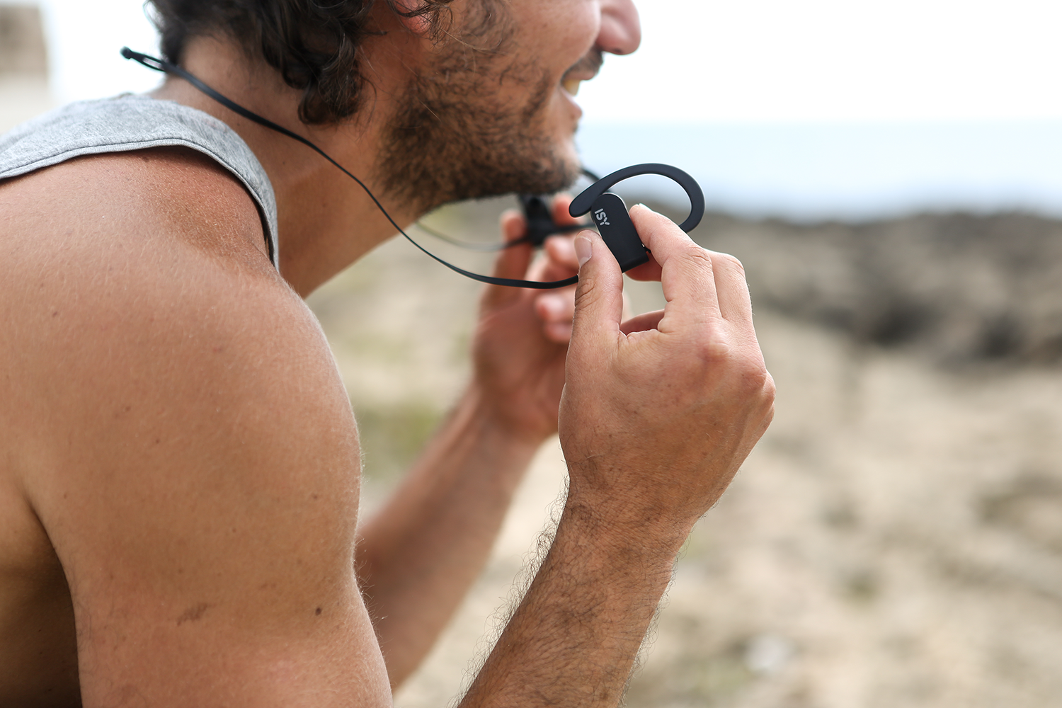 Man holding black wireless earphones. He is smiling. Outdoors, with a blurred background.