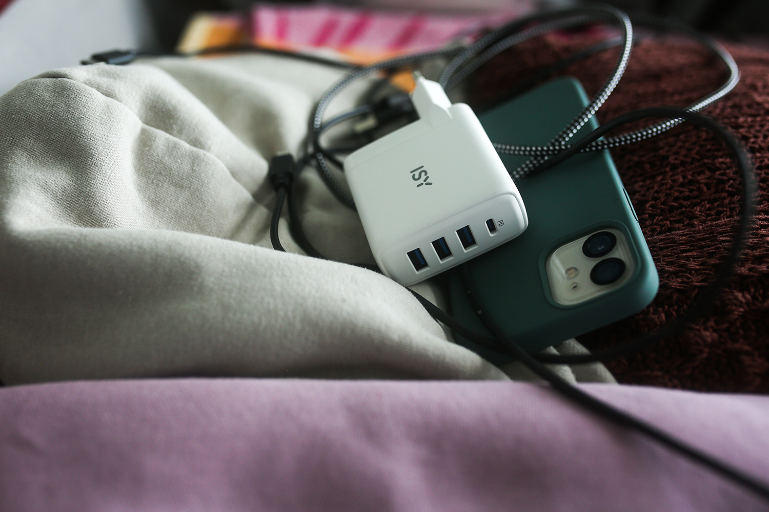 A white charger, a phone and a cable, on a beige bag and a pink blanket.