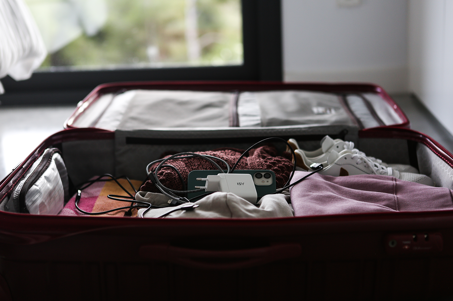 An open red suitcase filled with clothes, a phone, a charger and shoes, ready for travel.