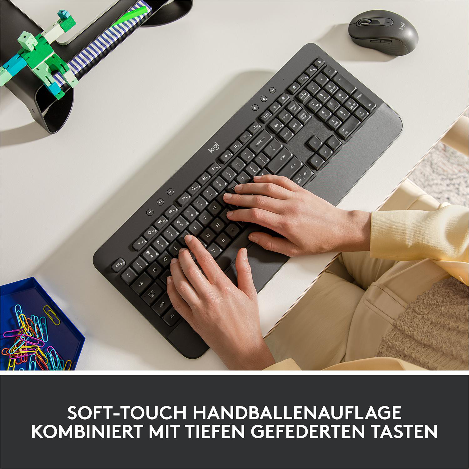 Eine Person tippt auf einer dunkelgrauen Tastatur. Eine Maus und einige bunte Büroklammern liegen auf dem Schreibtisch.
