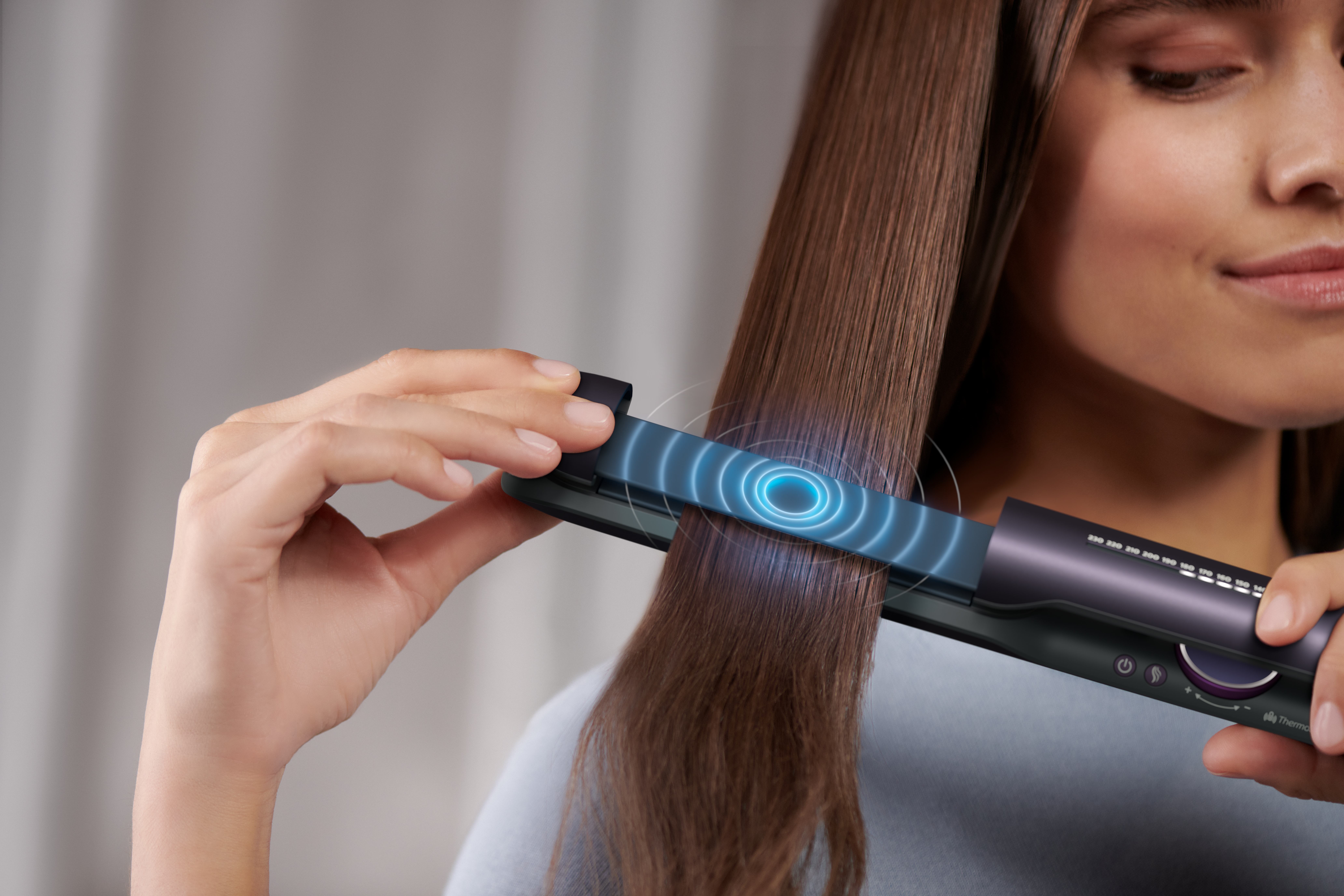 Una mujer usa una plancha de pelo. La herramienta está en el cabello, brillando azul. Ella sostiene la plancha con una mano.