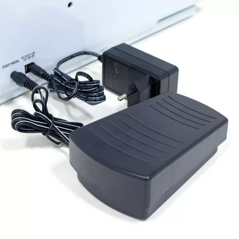 Lábpedál és hálózati adapter fehér felületen. Fekete pedál az előtérben.