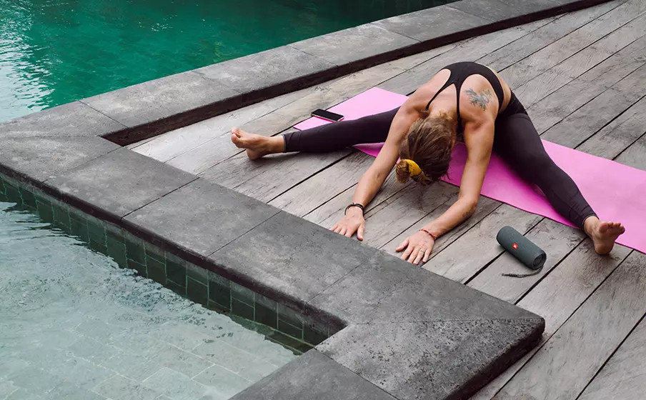Femme faisant du yoga près d'une piscine. Elle est sur un tapis rose, jambes écartées, touchant ses pieds.
