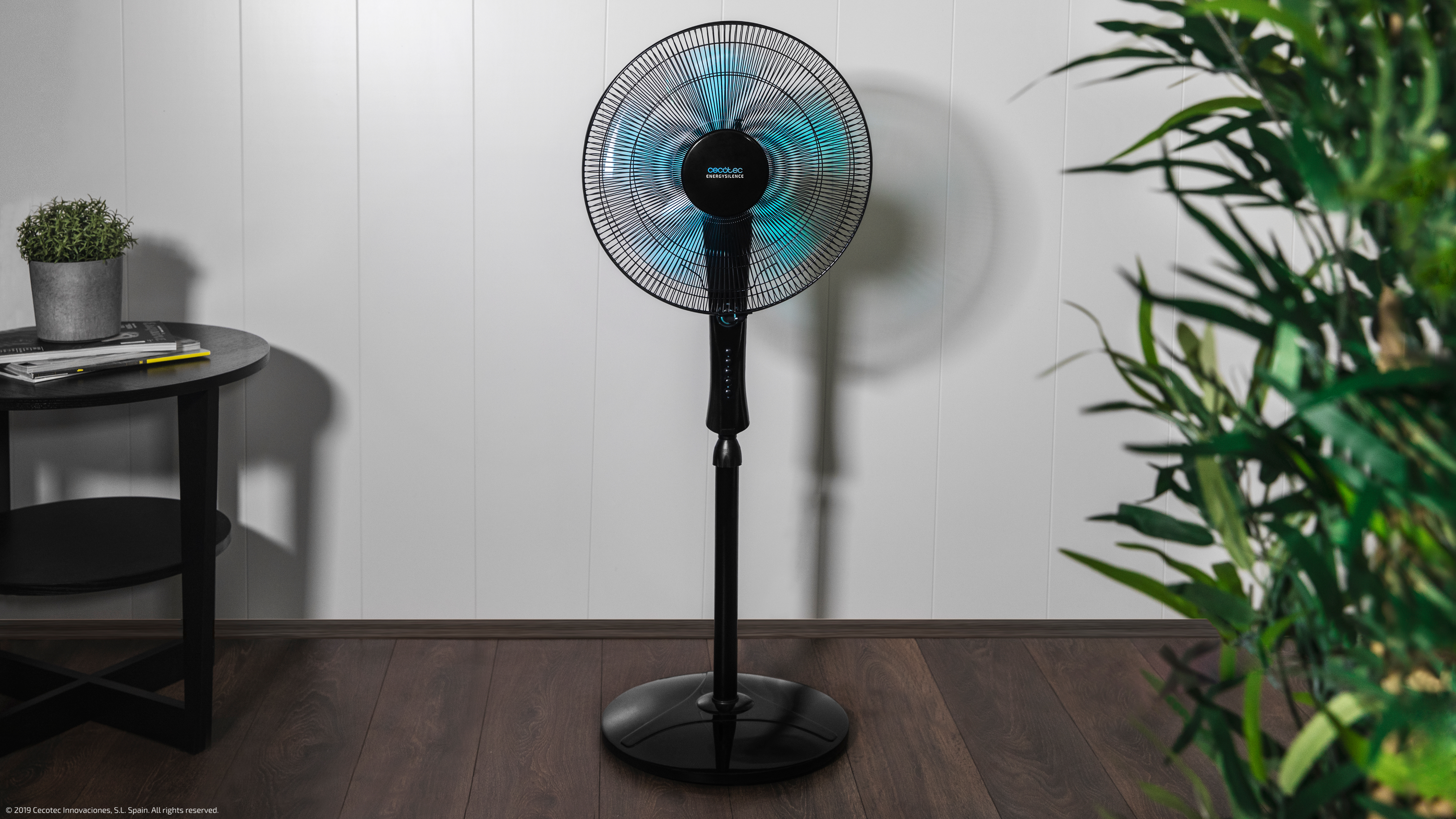 Un ventilador de pie negro en un suelo de madera, pared blanca.