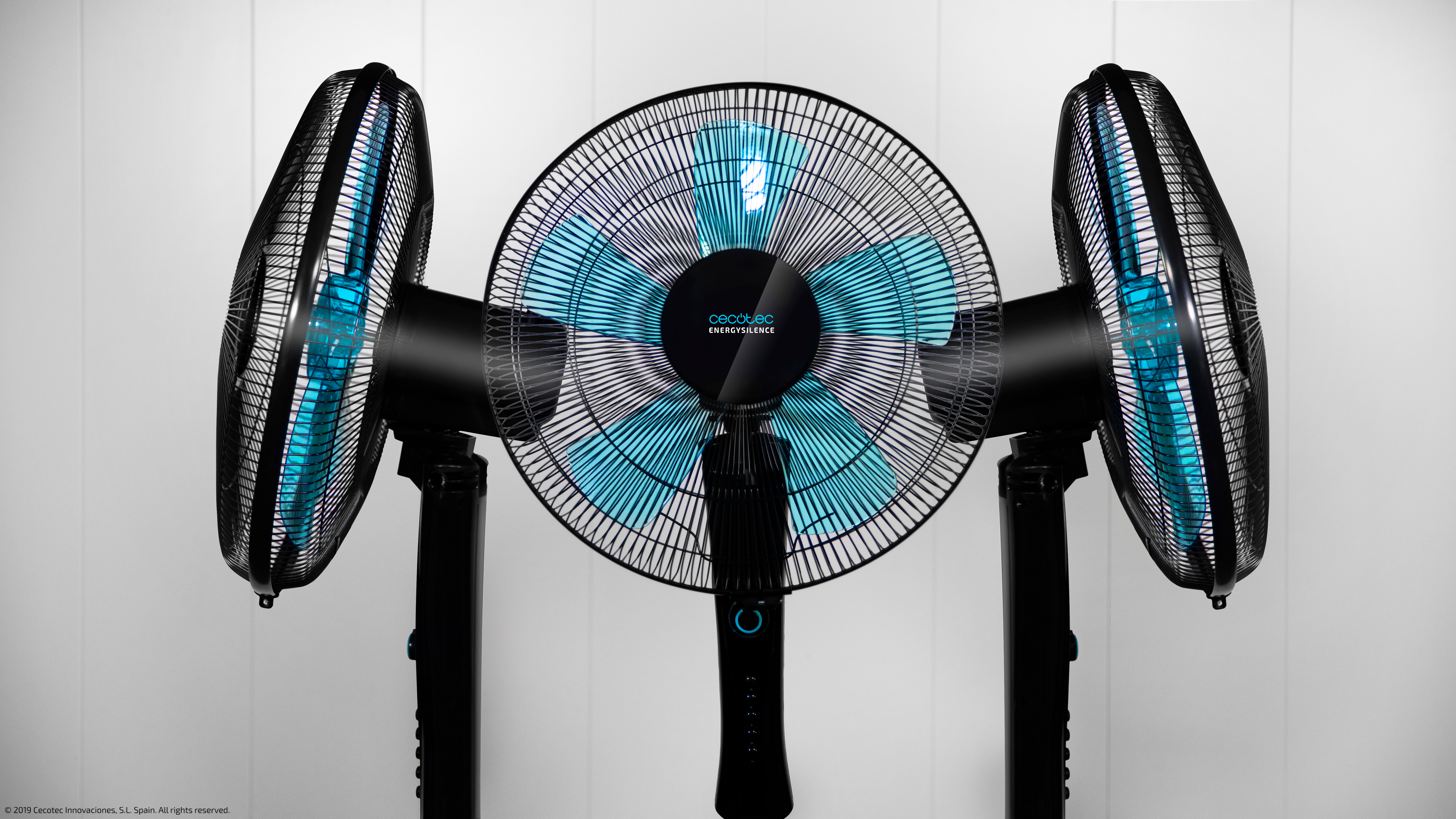Tres ventiladores negros sobre soportes con aspas azules contra un fondo de pared blanca.