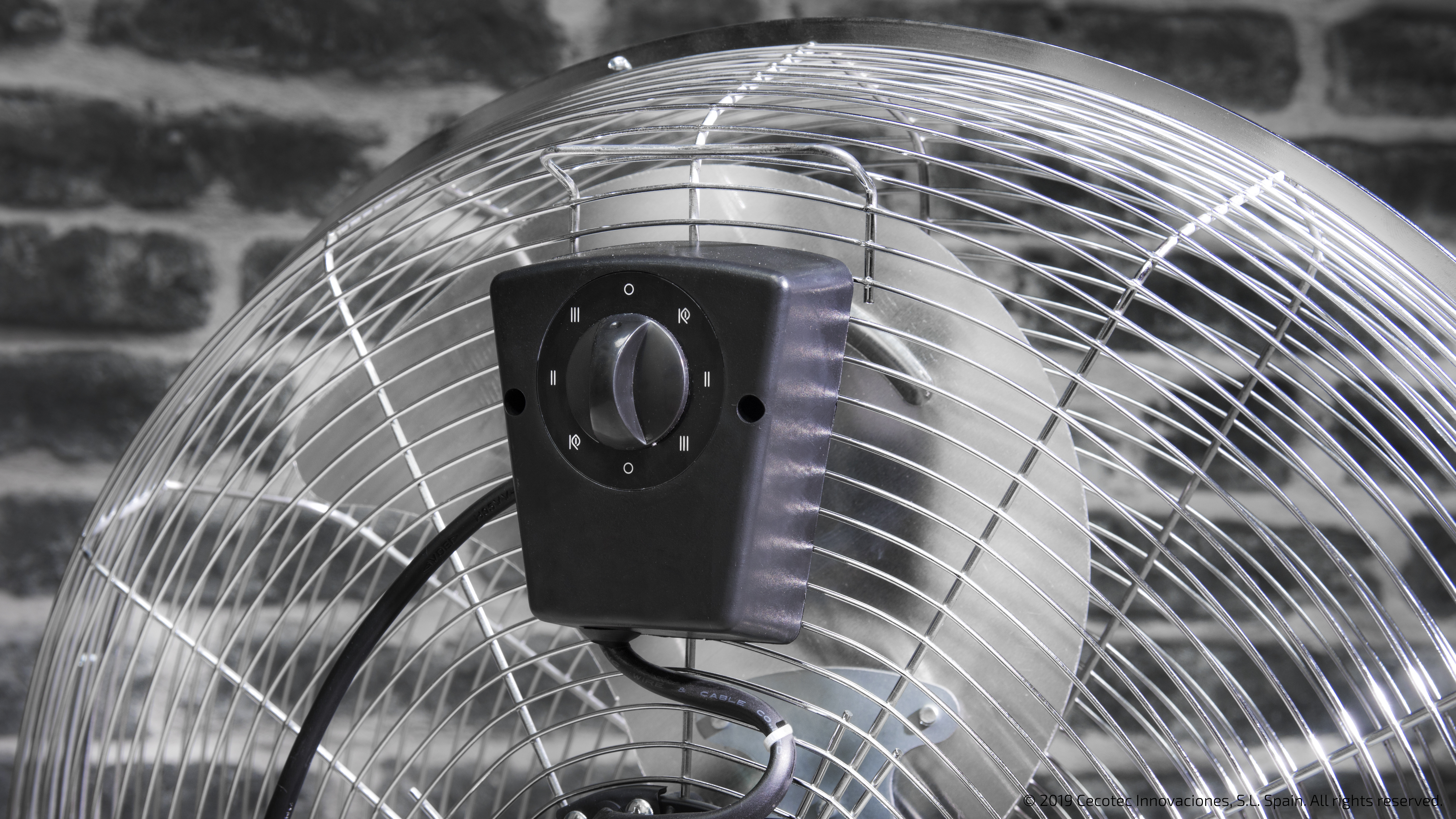 Primer plano de un ventilador plateado con un panel de control negro contra una pared de ladrillos.