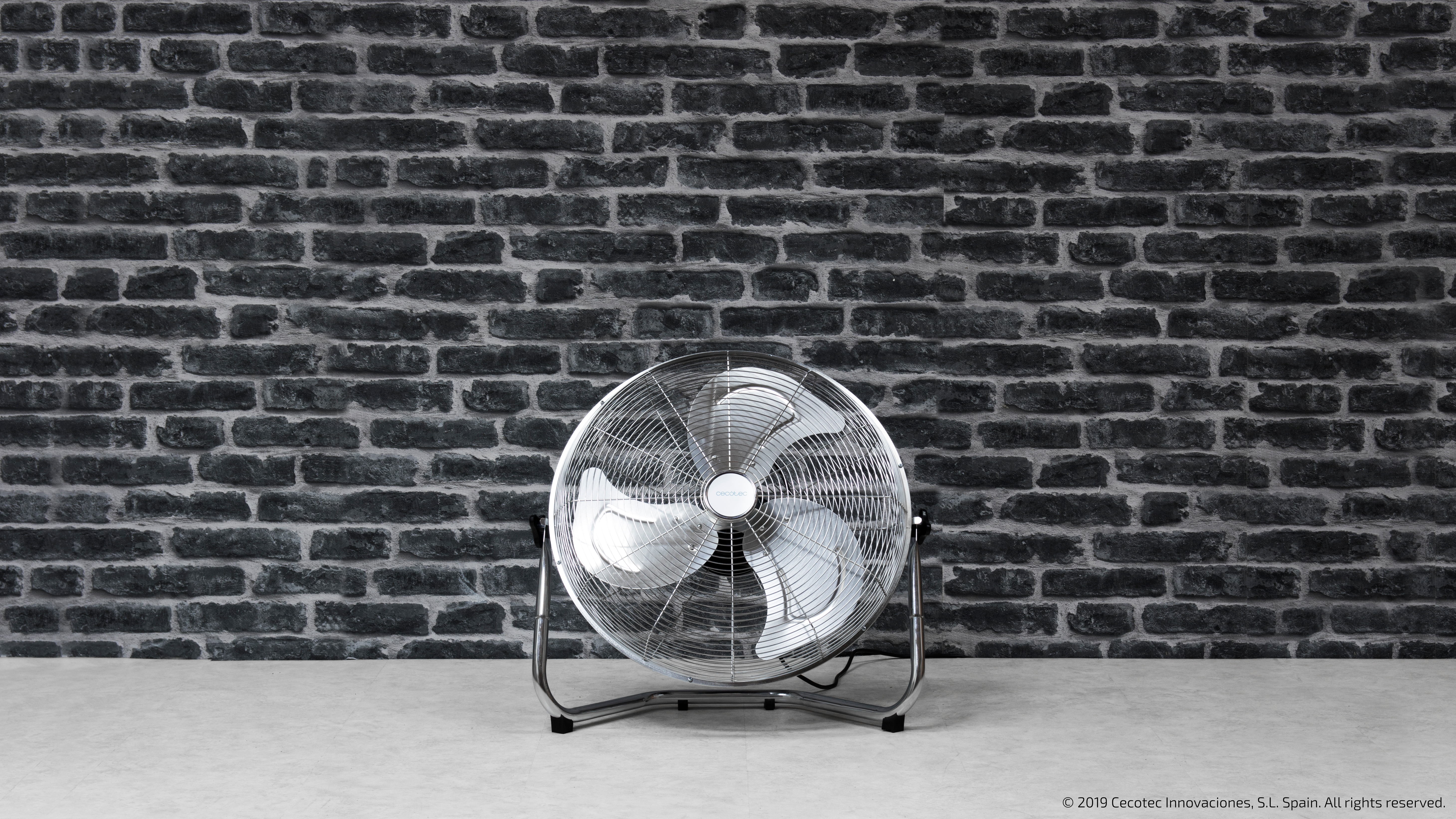 Ventilador plateado con aspas blancas frente a pared de ladrillos oscuros. Ventilador.