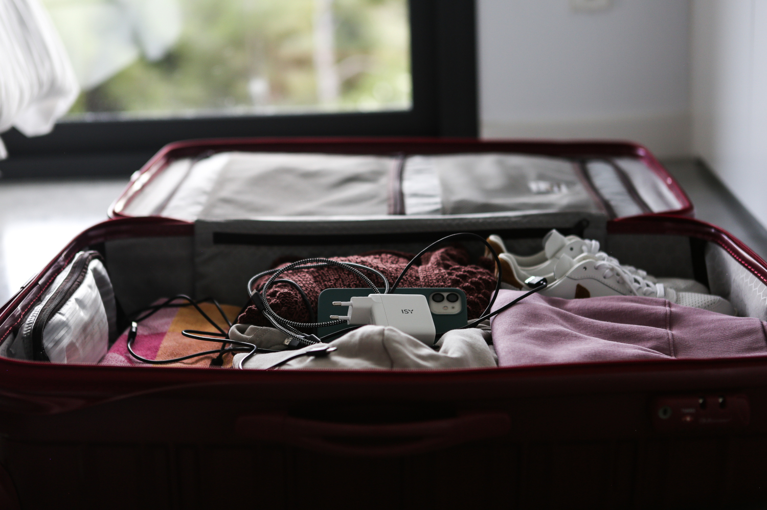 Een open koffer met kleding, oplader, telefoon en schoenen erin. De koffer is rood.
