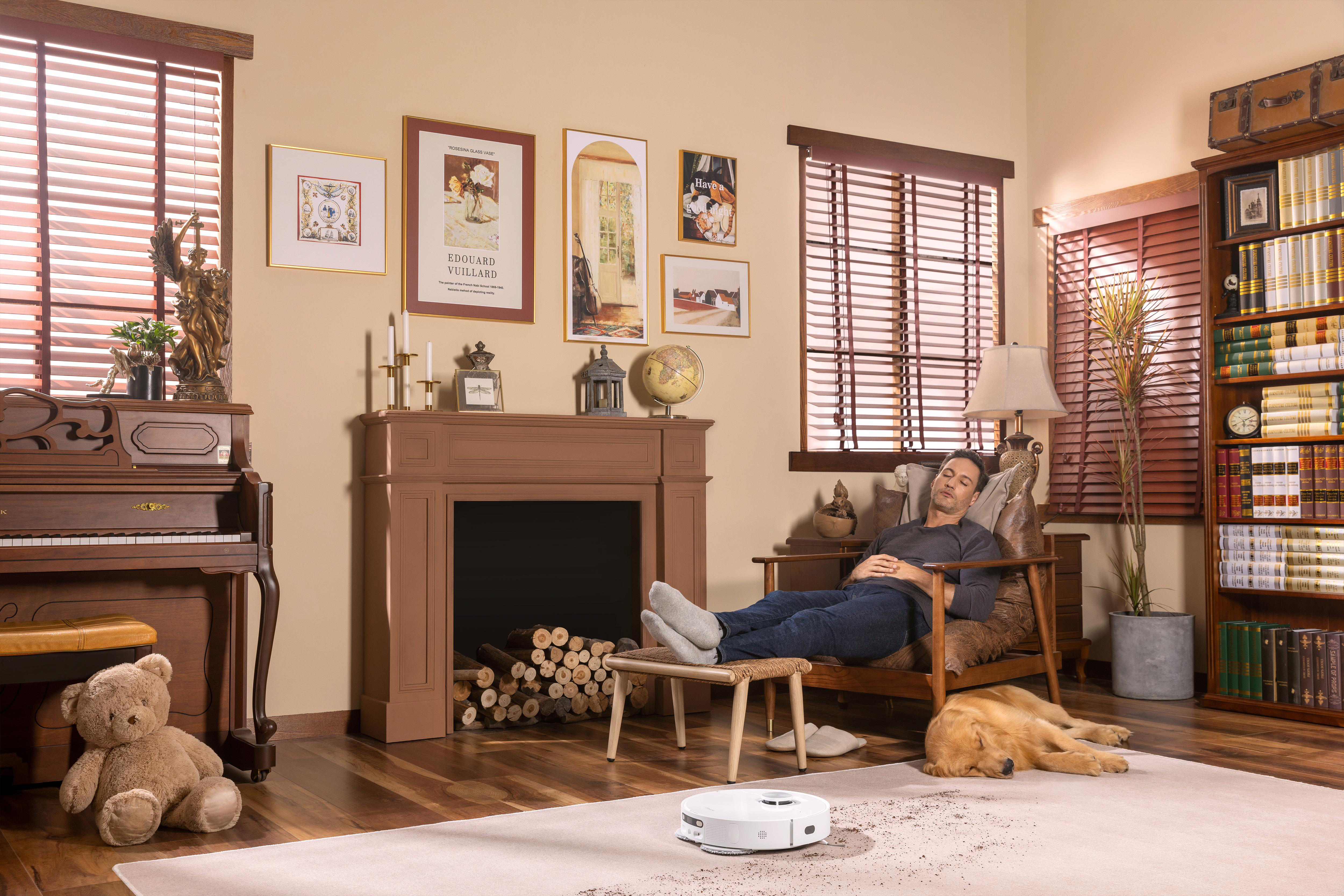 Mann entspannt sich in einem Wohnzimmer, mit Hund, Klavier, Gemälden, Kamin und Roboterstaubsauger.