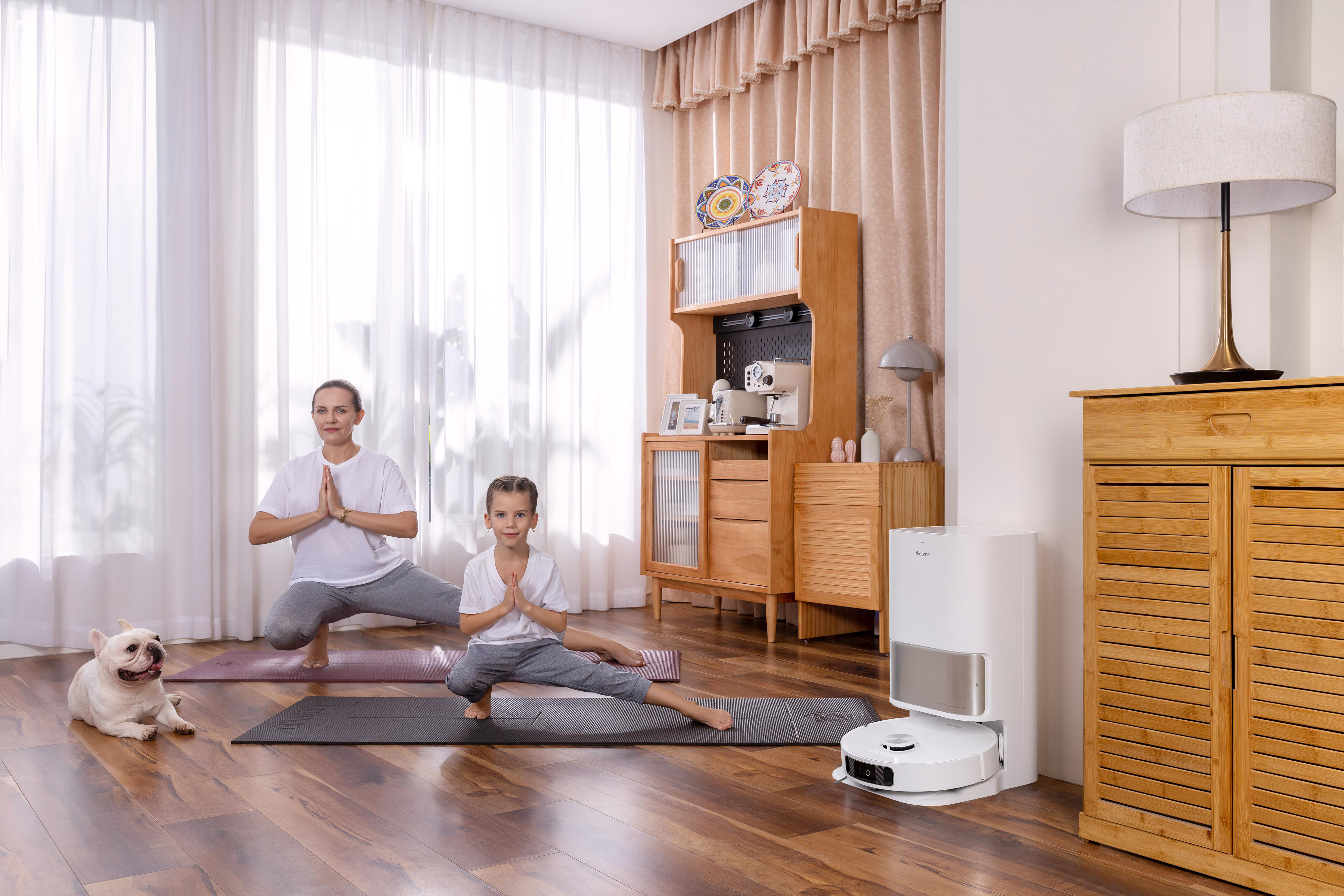 Frau und Mädchen in Yoga-Posen mit Hund. Ein weißer Roboterstaubsauger im Hintergrund.