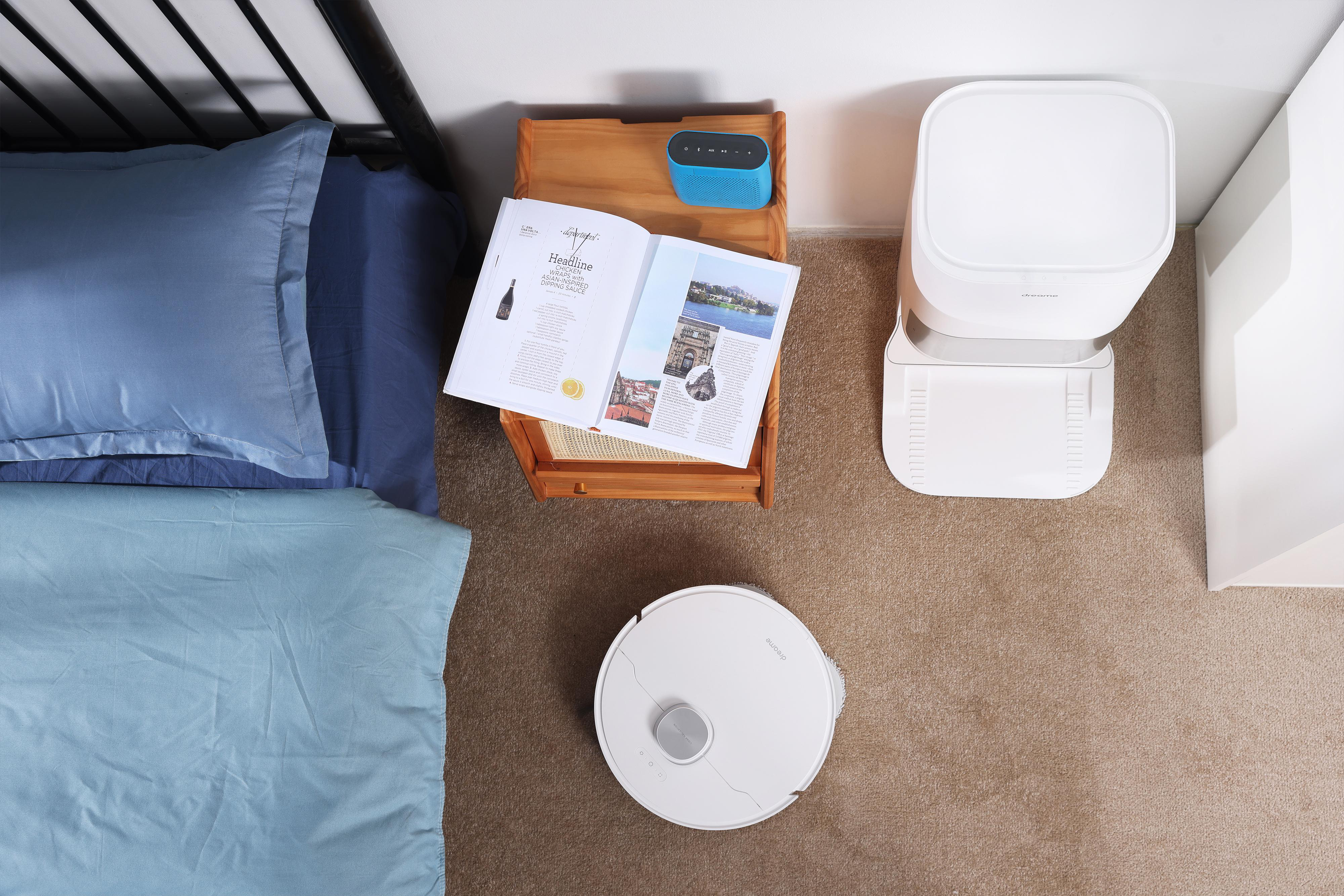 Draufsicht auf Schlafzimmer mit Bett, Tisch mit Buch und Lautsprecher sowie Roboter-Staubsauger.