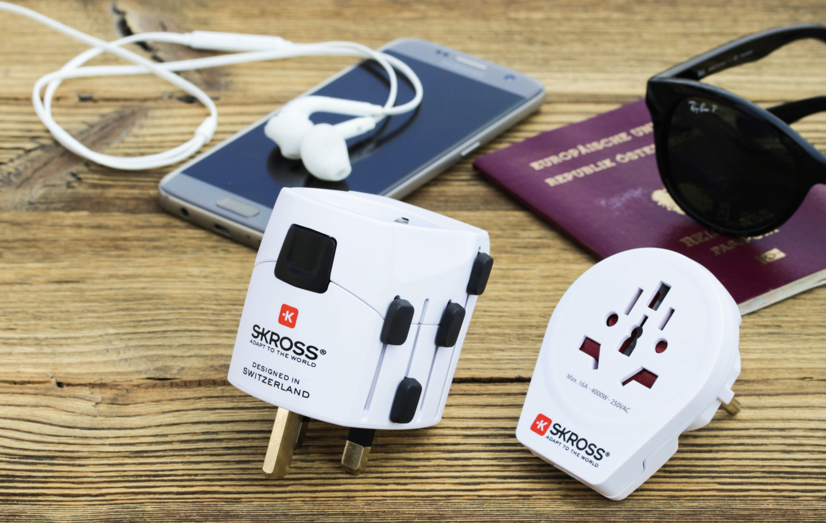 Un tavolo di legno con accessori da viaggio: un telefono, auricolari, un passaporto, occhiali da sole e due adattatori da viaggio bianchi.
