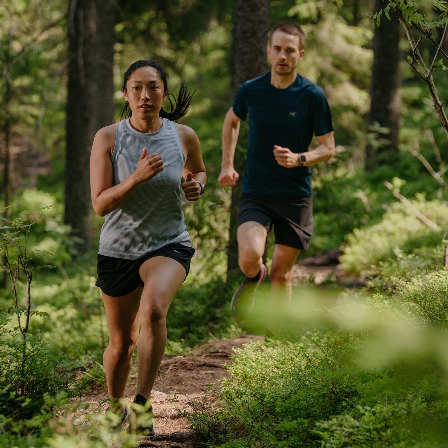 Dos personas corriendo por un sendero en un bosque. Árboles y arbustos verdes los rodean.