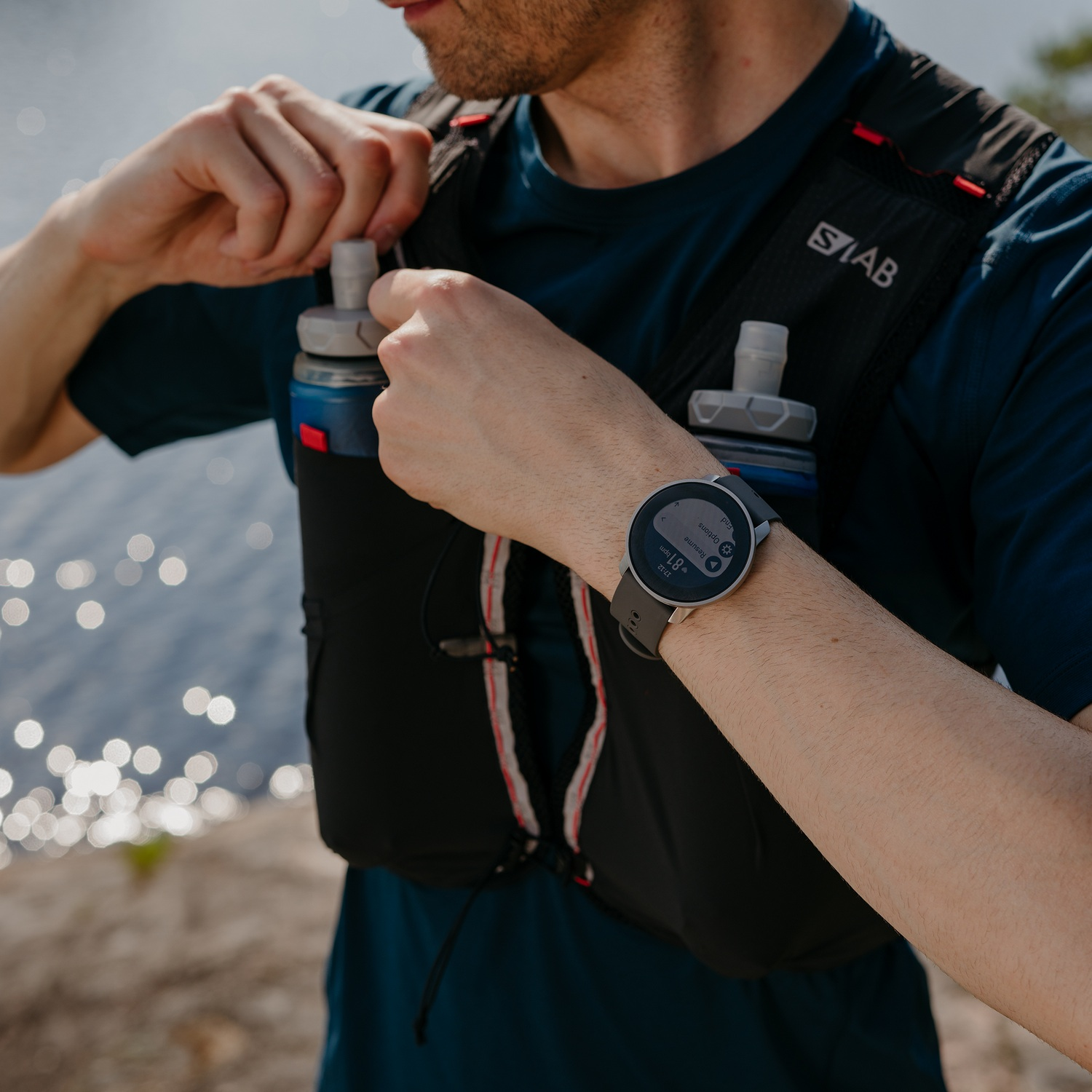 Hombre con chaleco y reloj de running, llenando una botella de agua. El fondo es un lago.